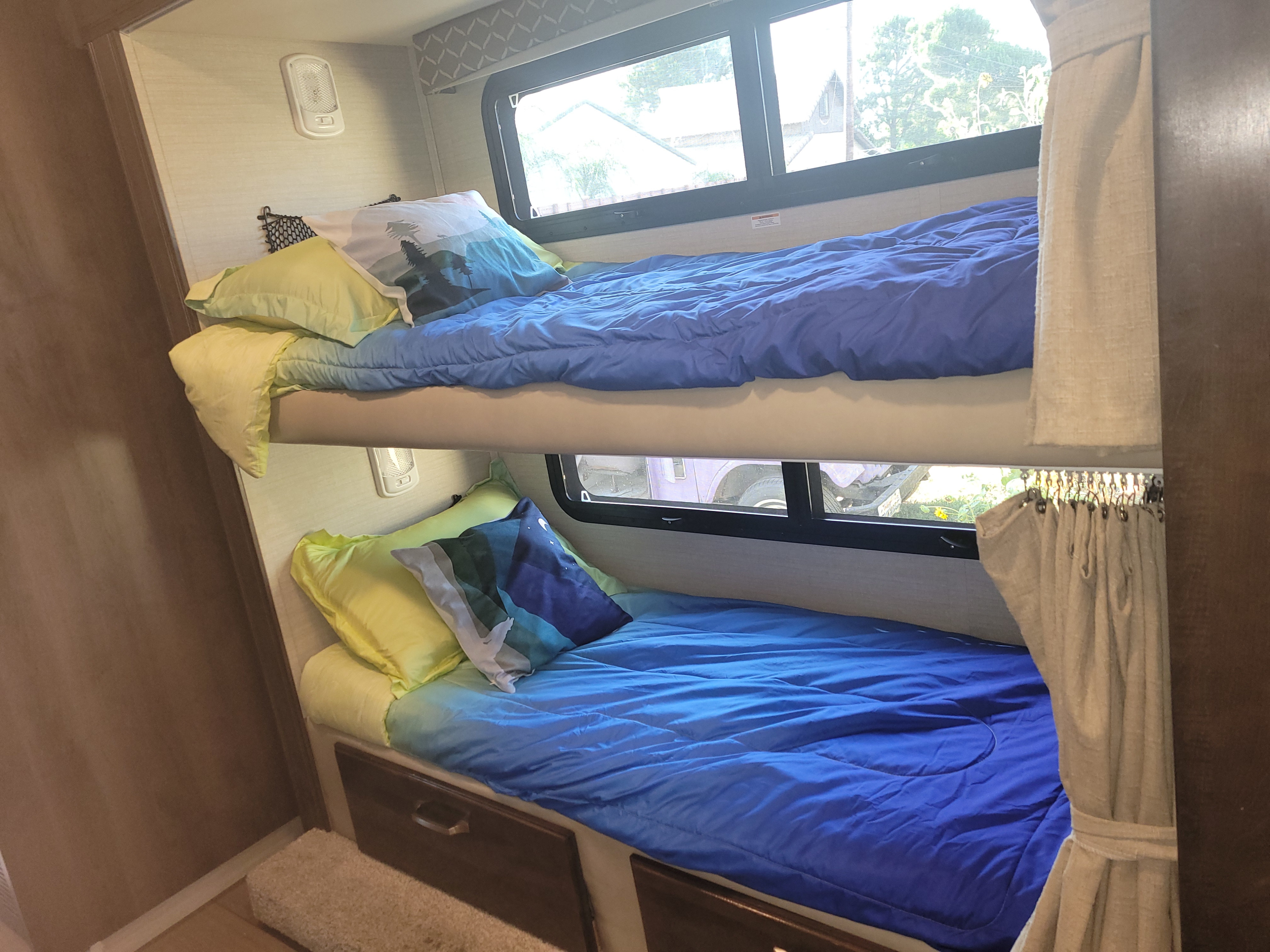 Inside an RV rental, two bunk beds with blue comforters & decorative pillows offer comfy sleeping, with windows & storage drawers visible.. Jayco Greyhawk 2018