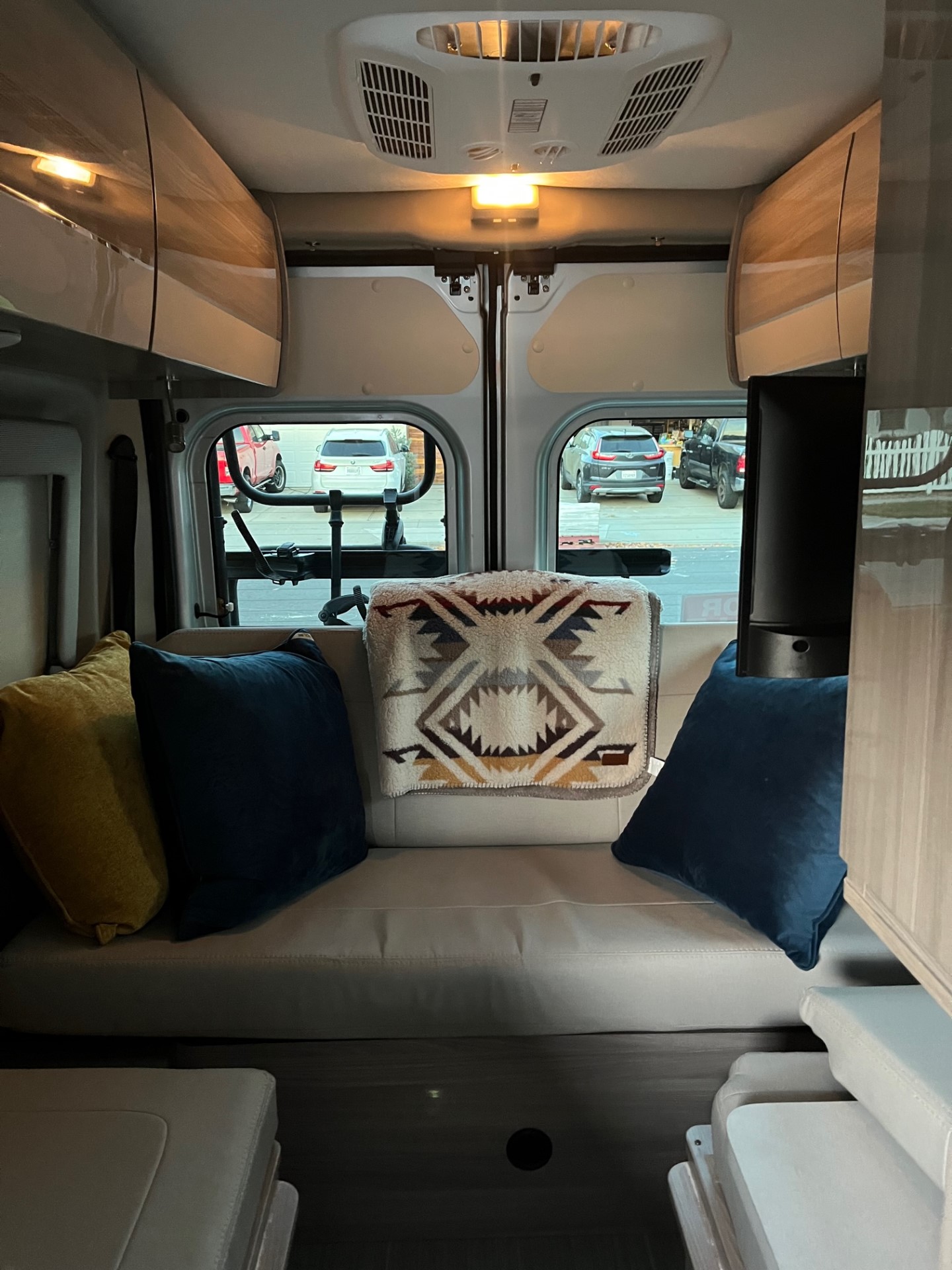 View from an RV rental: cozy interior with cushions and a blanket, looking out at parked vehicles.. Thor Sequnce 2023