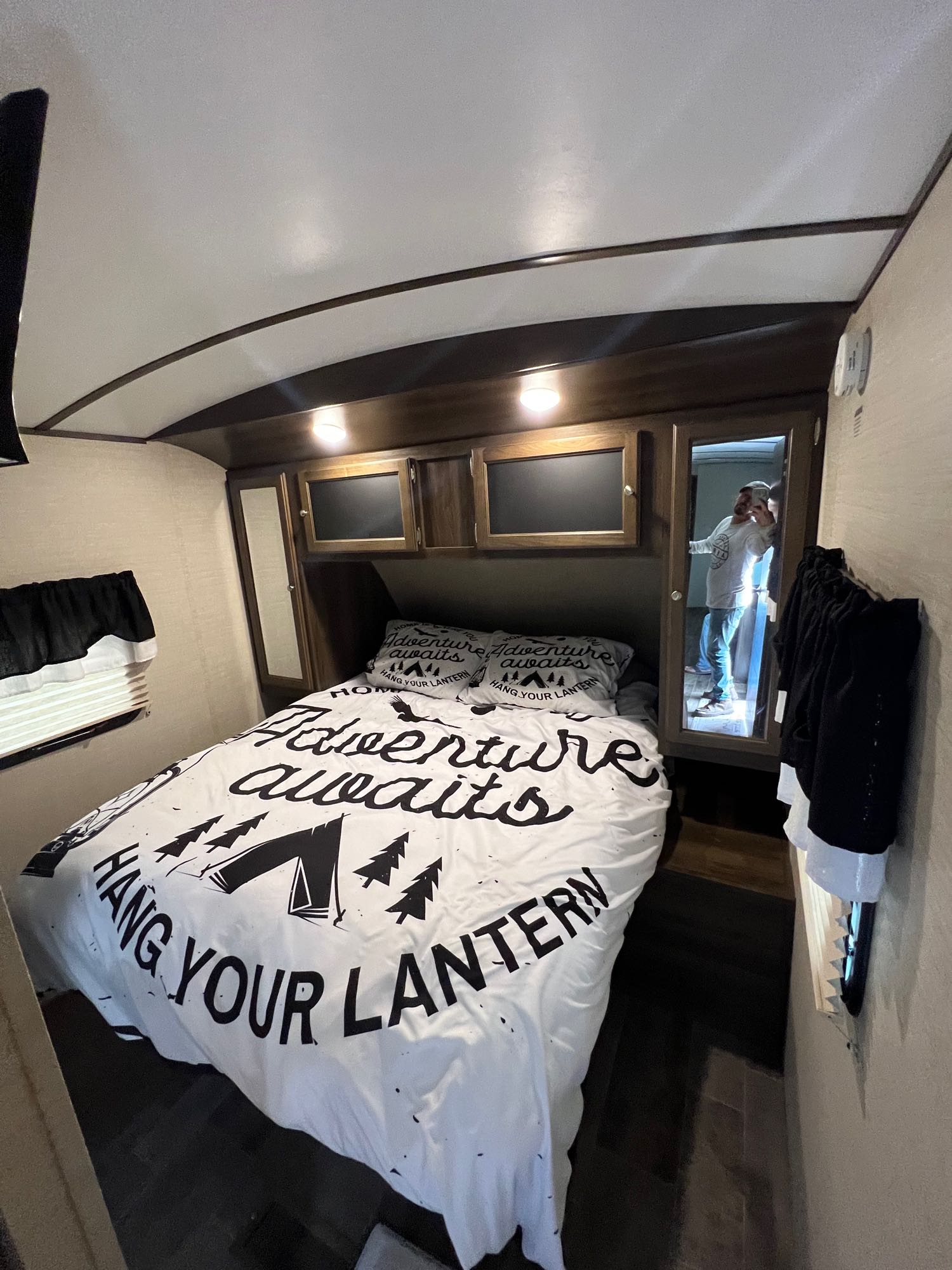 View inside an RV rental bedroom: bed with adventure-themed duvet, wood cabinets, and mirror reflecting a person.. Keystone RV Bullet Premier 2018