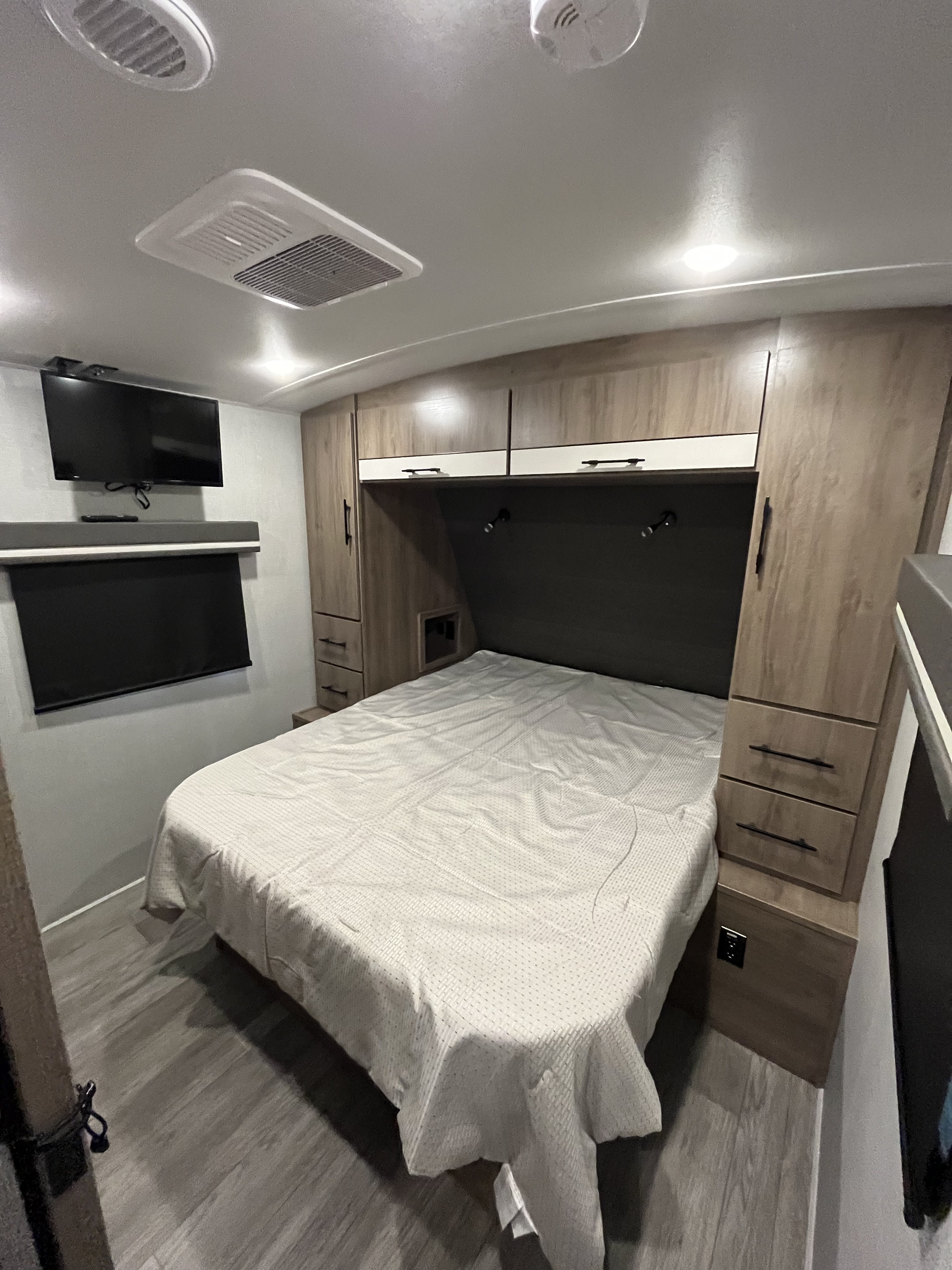 Compact RV rental bedroom with a neatly made bed, wall-mounted TV, wood-paneled storage, and ceiling vents.. Grand Design Imagine 2023