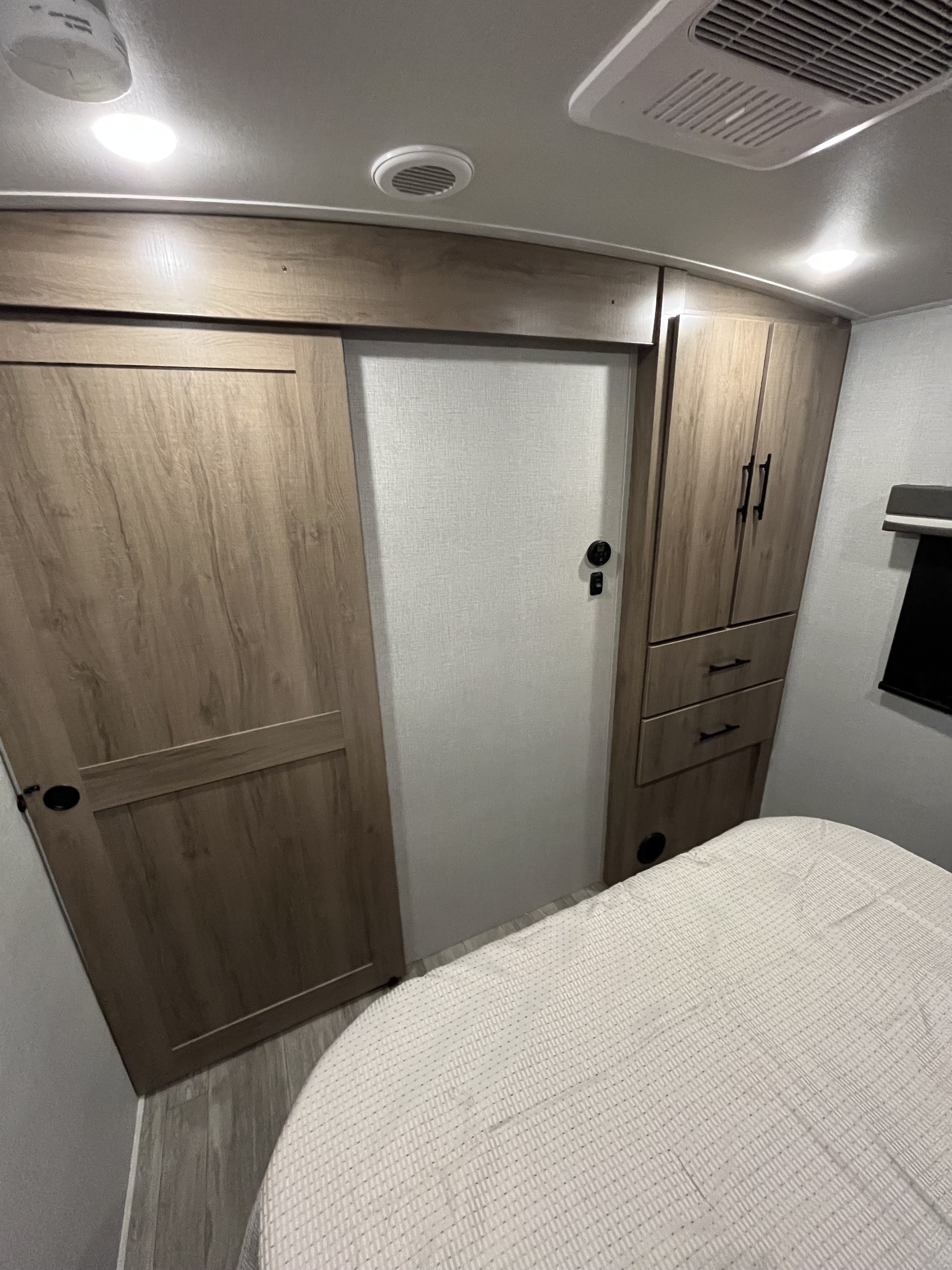 Modern RV rental interior showing a bedroom area with light wood storage, a sliding door, and a cozy bed.. Grand Design Imagine 2023
