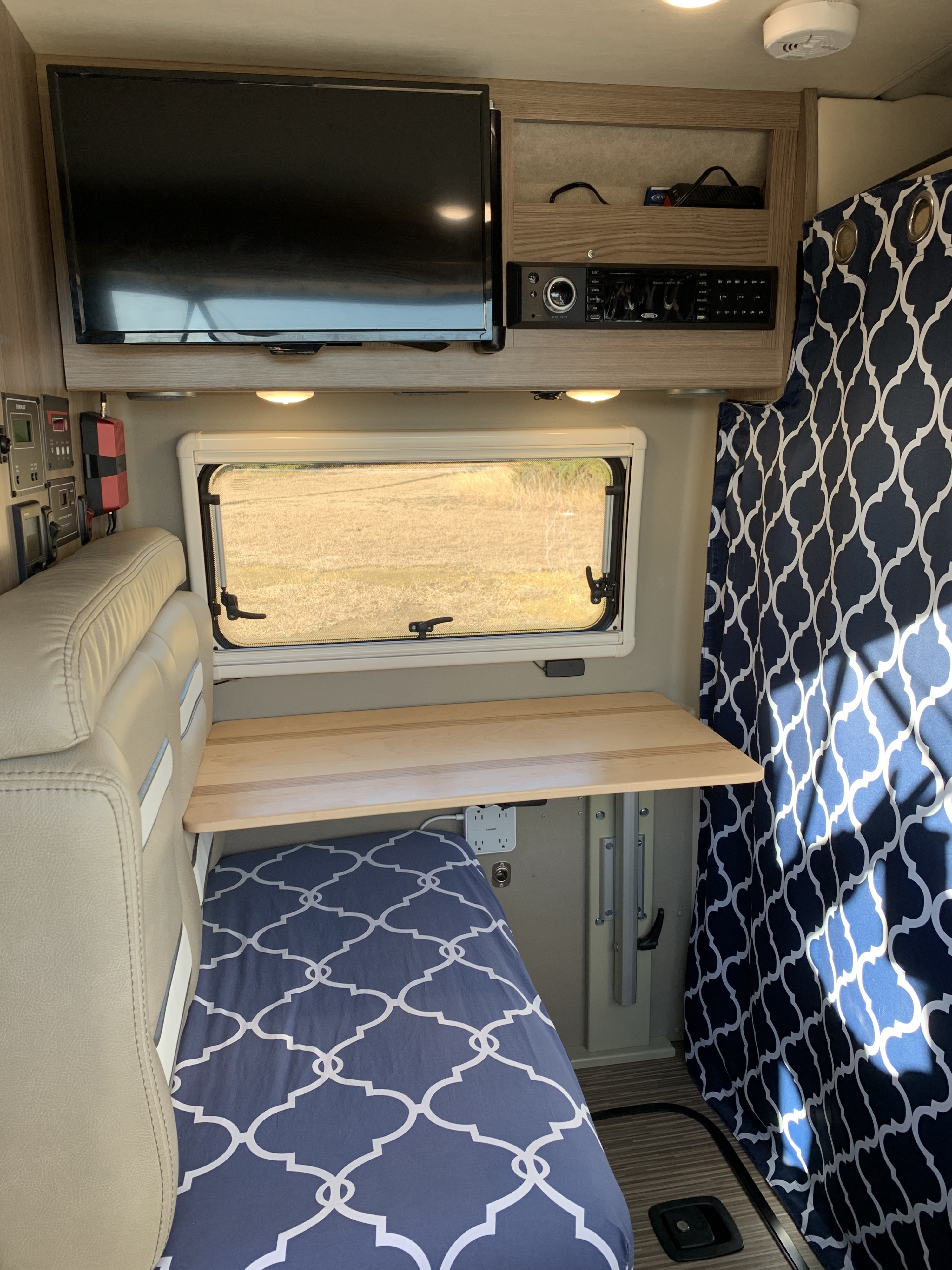 RV rental interior view: shows a TV, stereo, window, fold-out table, and blue patterned bedding & curtain.. Winnebago Travato 2020
