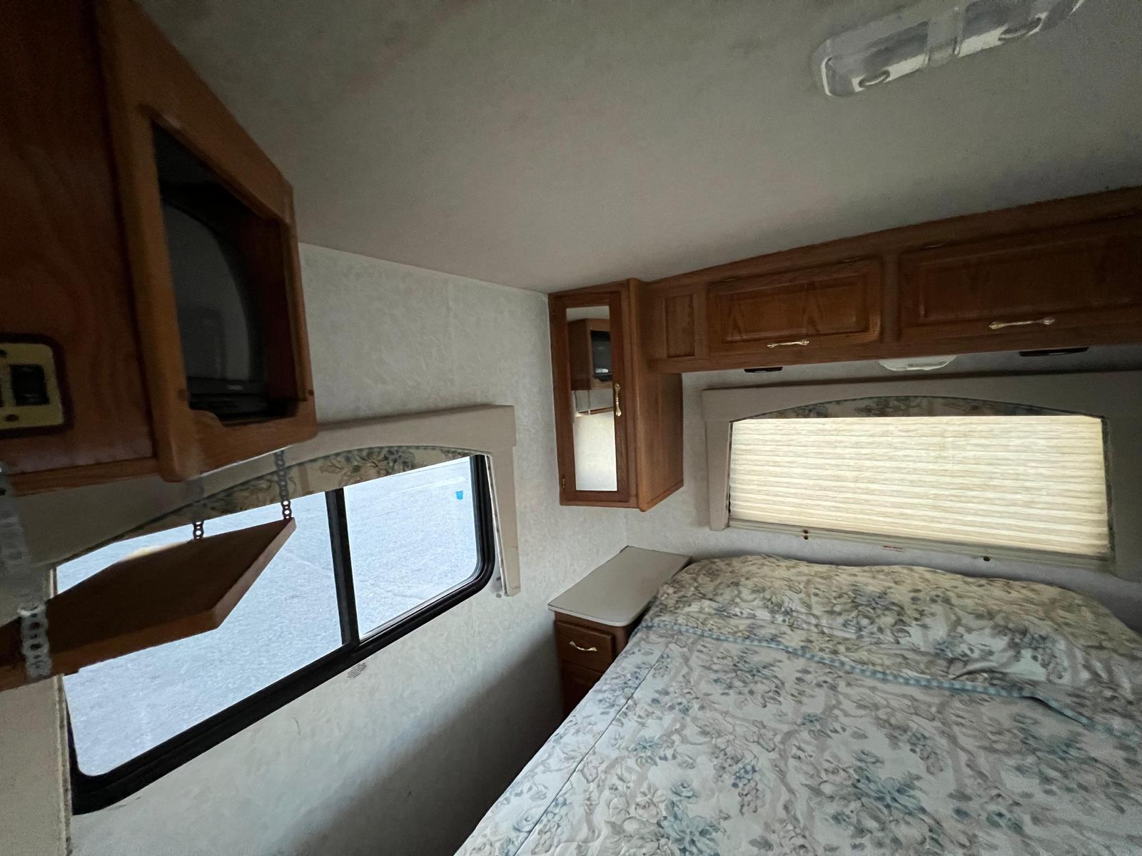 Cozy RV rental bedroom features a floral bed, overhead wood cabinets, and an open window, perfect for mobile travel.. Allegro Allegro Motorhome 2000