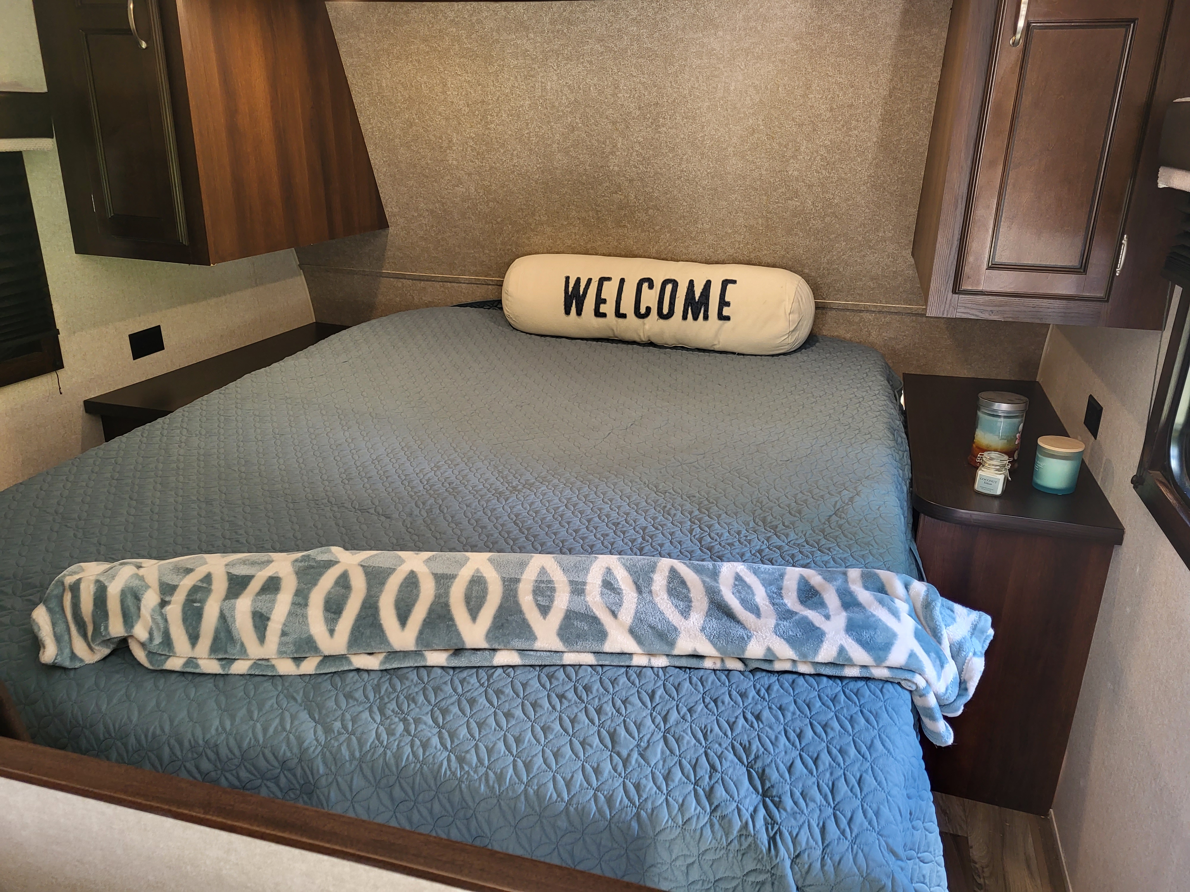 Cozy RV rental bedroom featuring a blue bed with a patterned blanket, "WELCOME" pillow, and bedside decor.. Jayco Jay Flight 2018