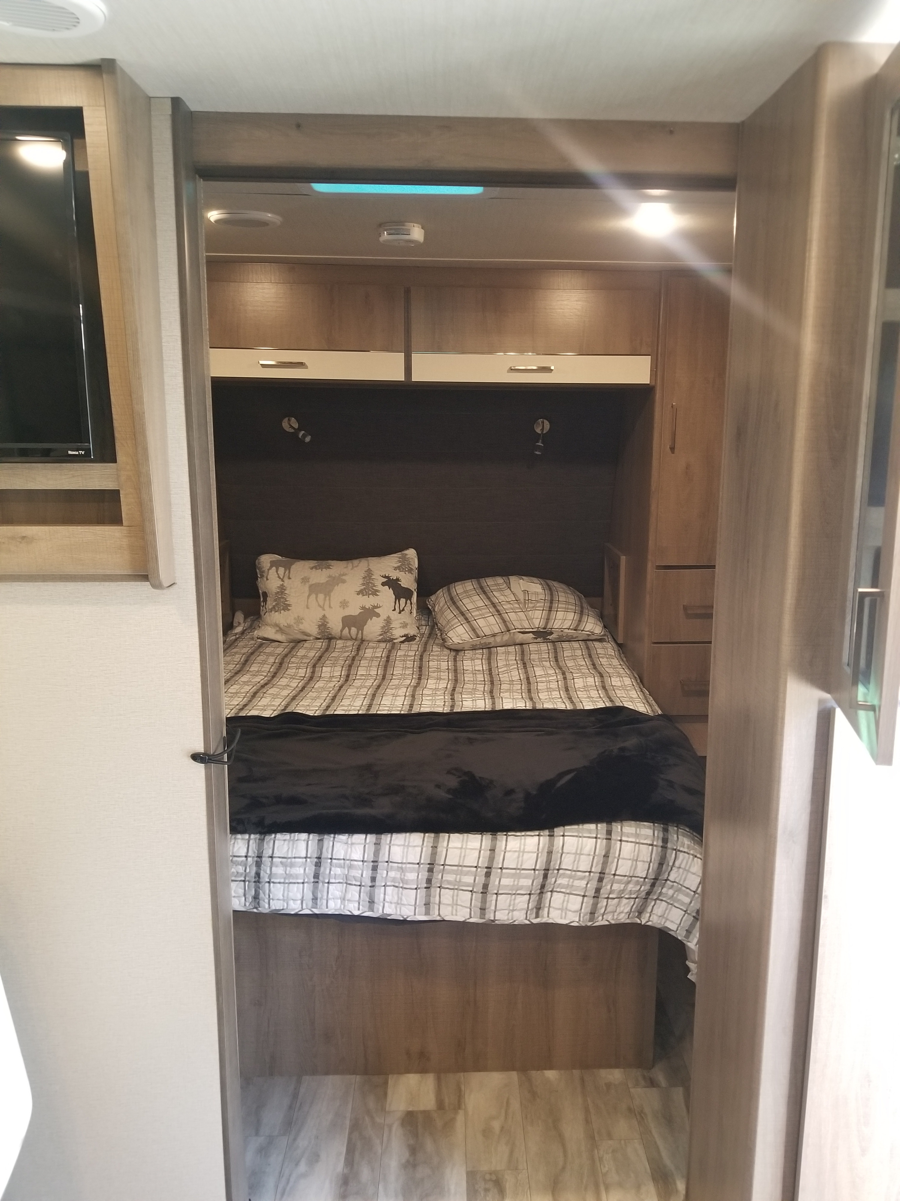 RV rental's cozy bedroom interior, featuring a plaid bed with moose pillow, overhead cabinets, reading lights, and side storage.. Grand Design Imagine XLS 2021