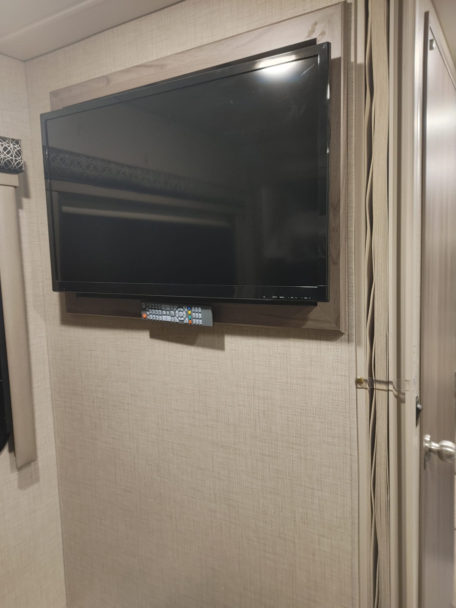 RV rental: Wall-mounted TV with a remote on a small shelf, integrated into a wood-framed section of a textured RV wall.. Thor Tiburon 2022