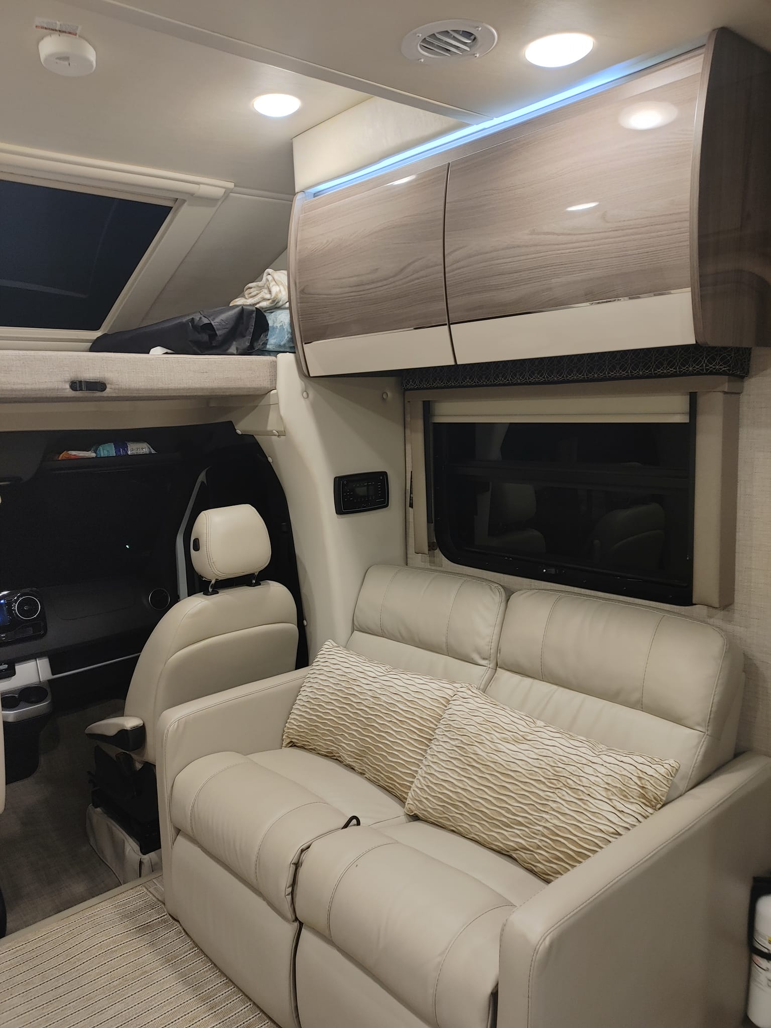 This RV rental interior shows a cream leather sofa, overhead cabinets, and a cab-over sleeping space with bright lighting.. Thor Tiburon 2022