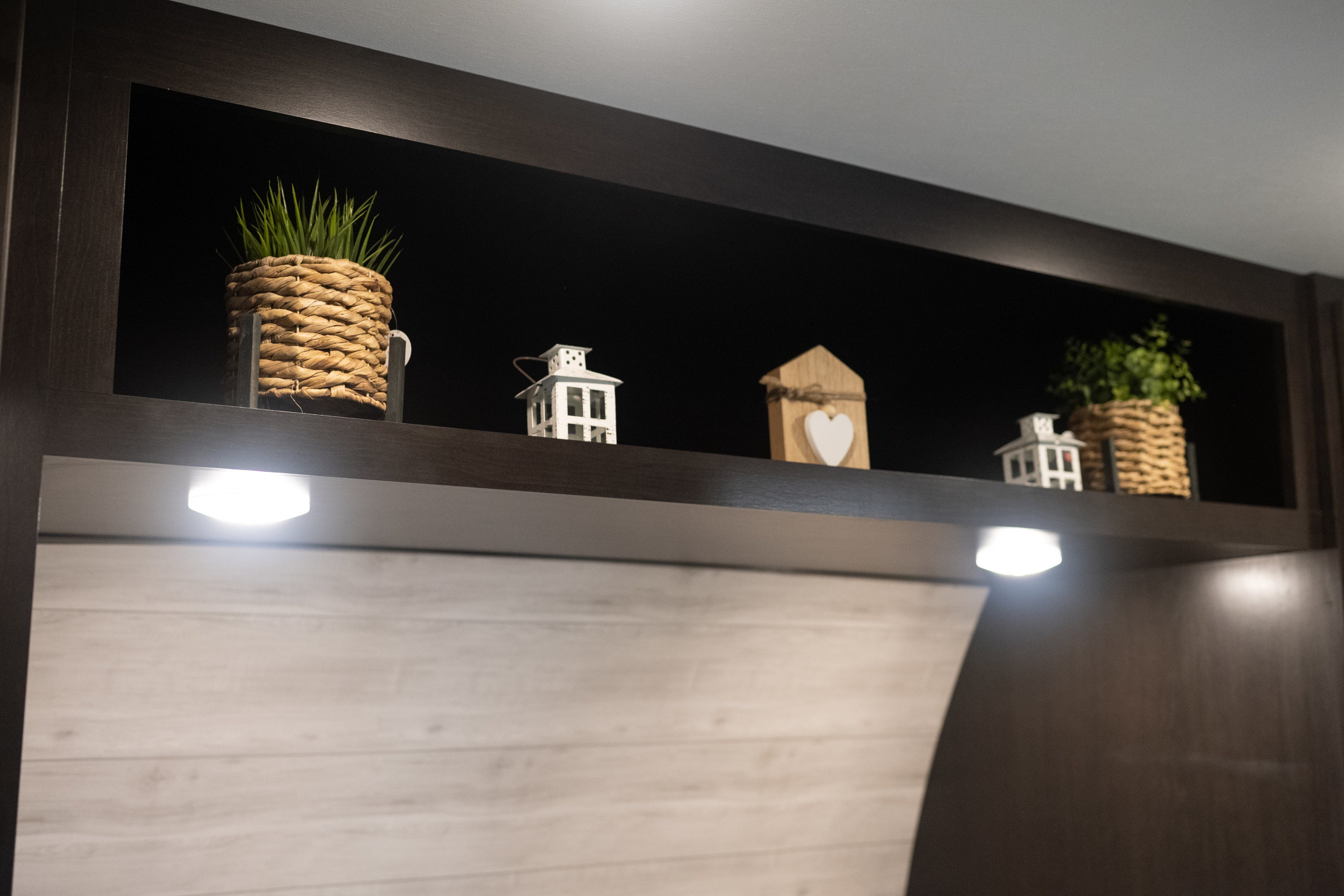 Overhead storage in an RV rental, decorated with green plants, lanterns, and a wooden house; illuminated by under-shelf lights.. Jayco Jay Flight 2023