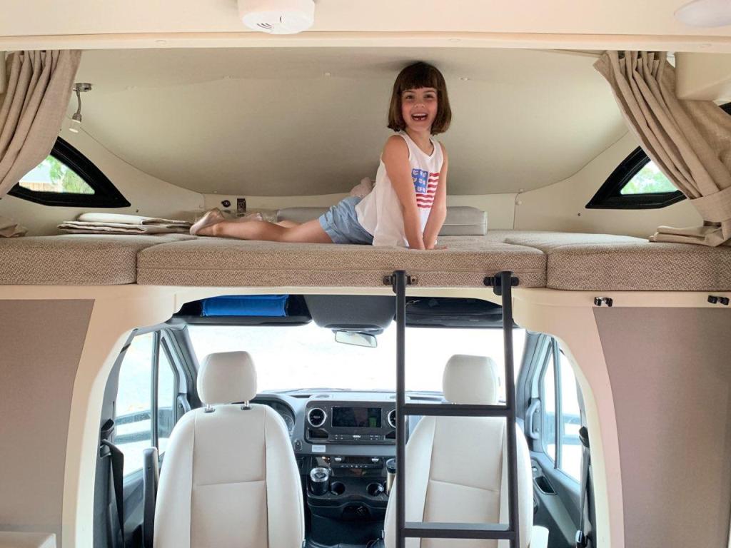 Playful girl smiles from the cozy overhead bunk in this RV rental. Below, the spacious driver's cabin completes the camper's interior.. Winnebago View 2023