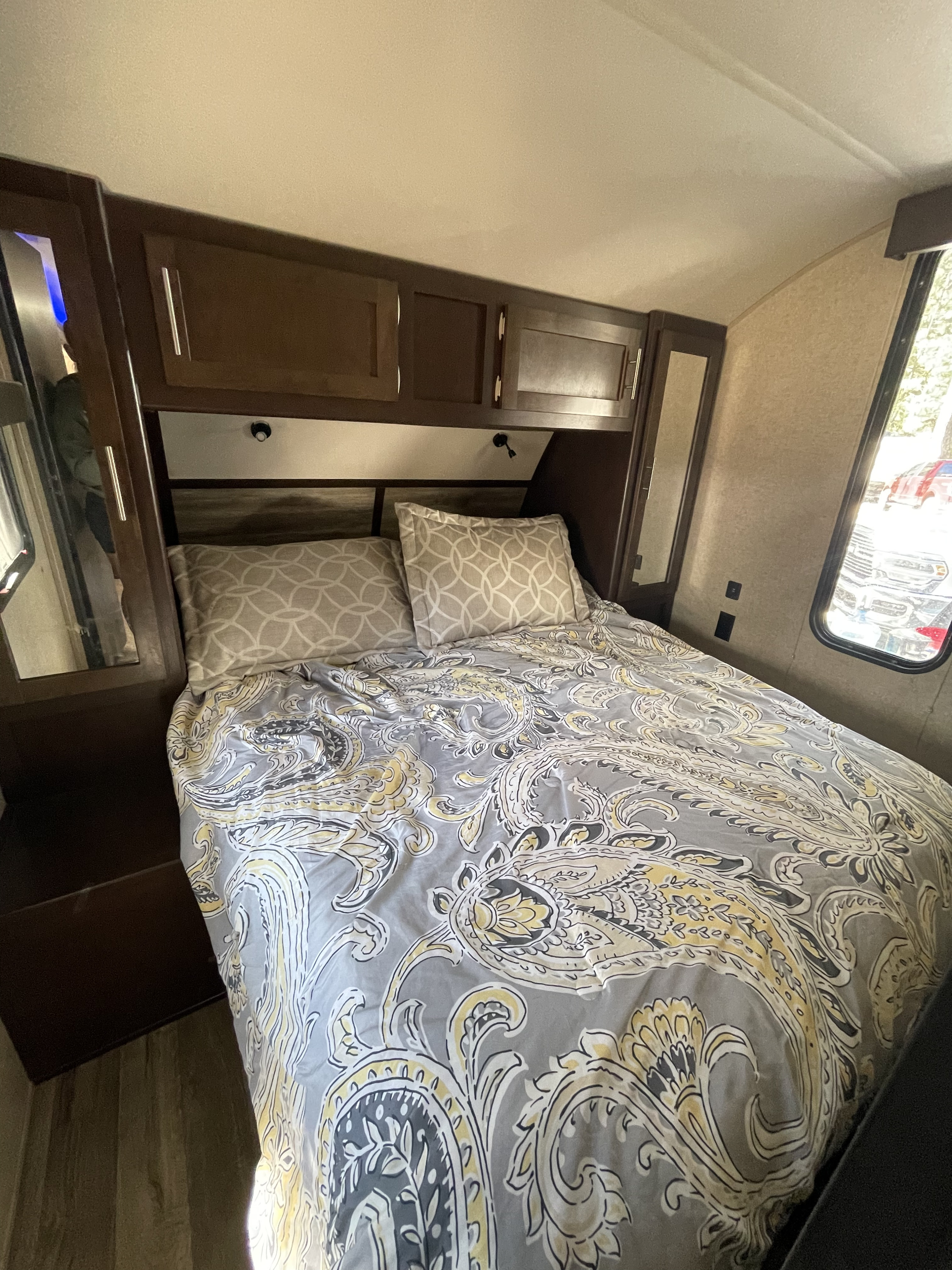 RV rental bedroom: cozy bed with grey/yellow paisley bedding, decorative pillows, and dark wood overhead cabinets.. Forest River Cherokee 2018