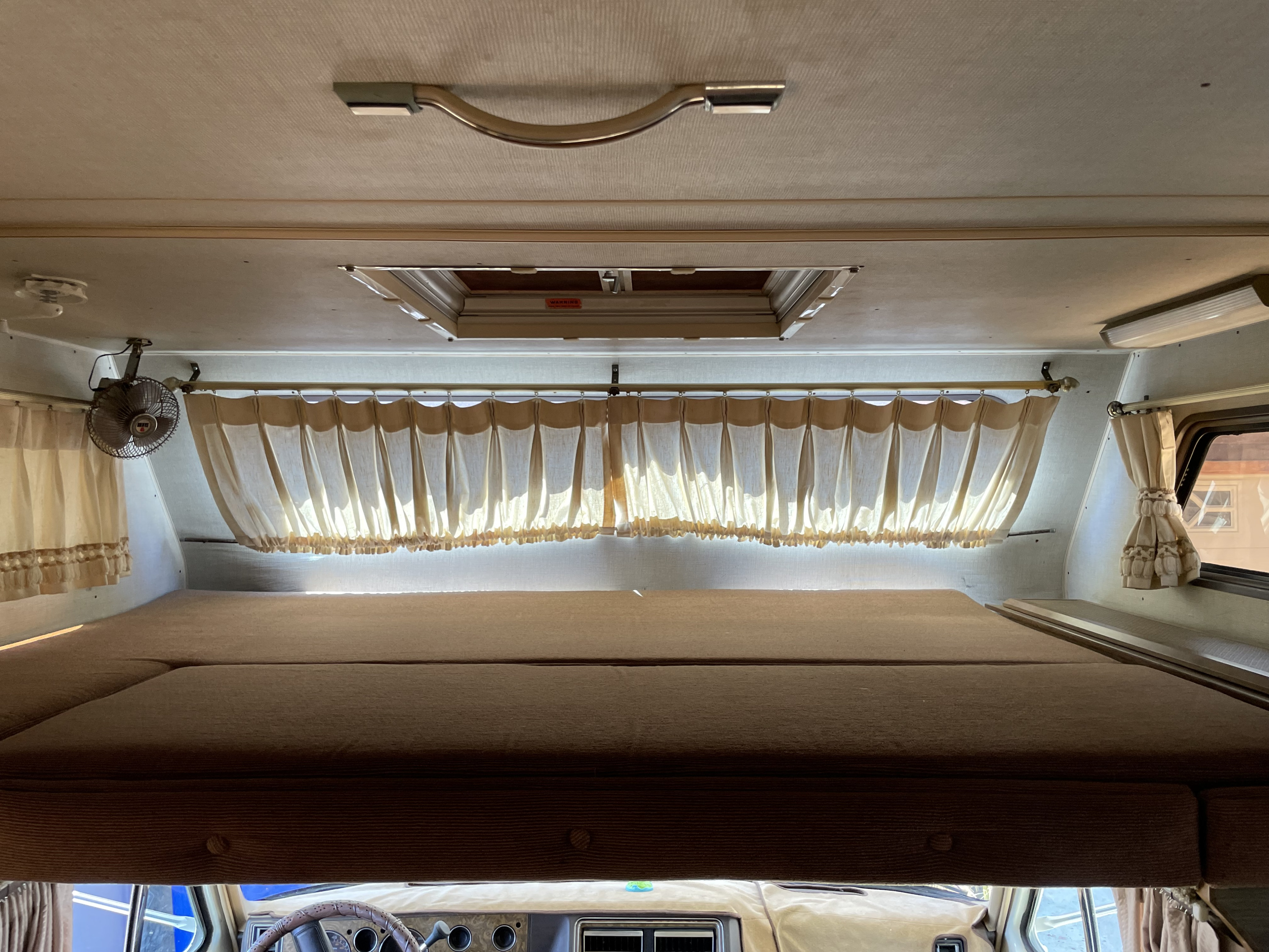 Looking into the cozy cab-over bunk area of an RV rental, showing curtains, a fan, and a roof vent.. Chevrolet Lazy Daze 1985