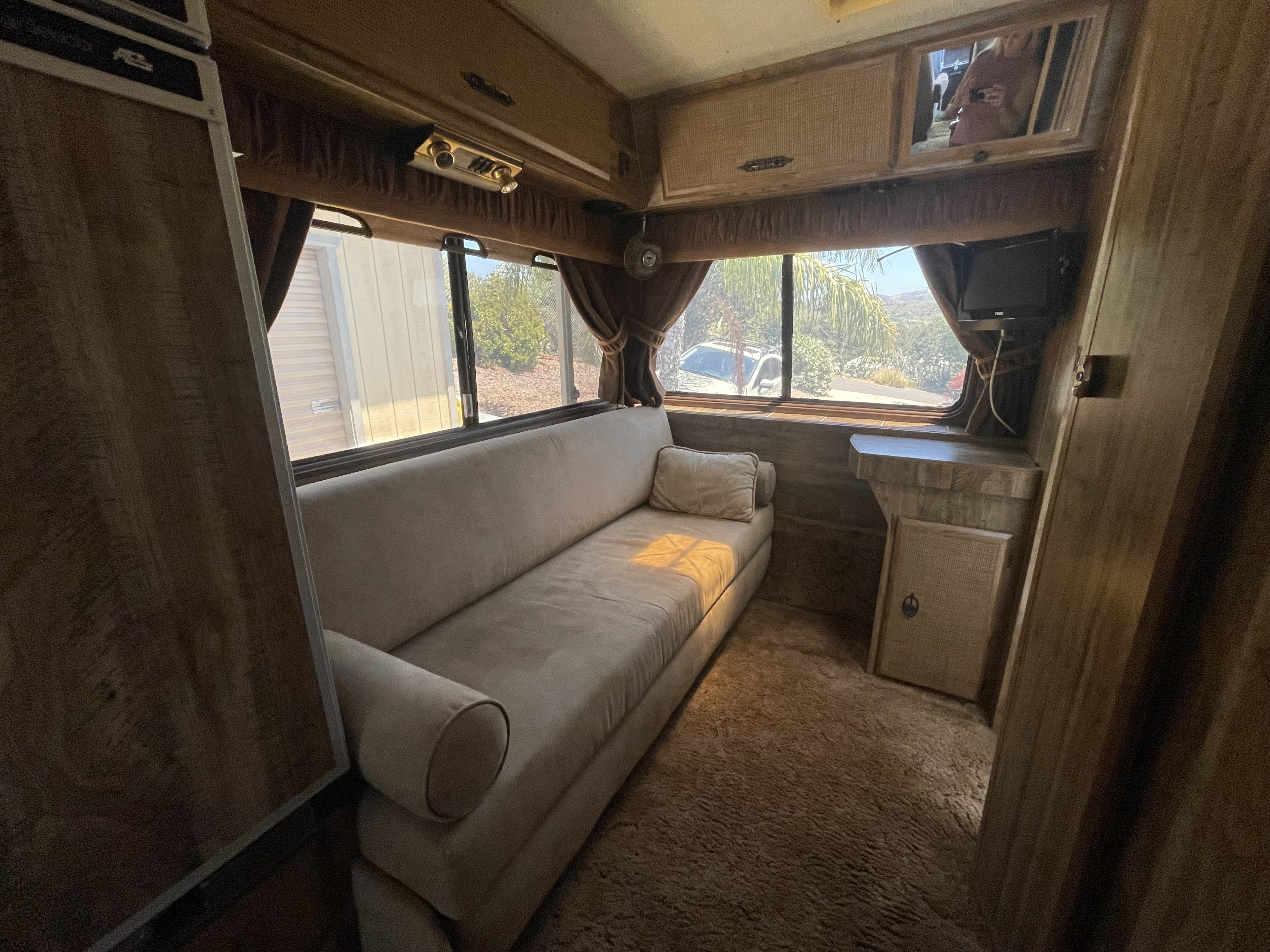 Inside an RV rental: A warm living space with a large, light-colored sofa, wood paneling, and views through expansive windows.. Chevrolet Lazy Daze 1985