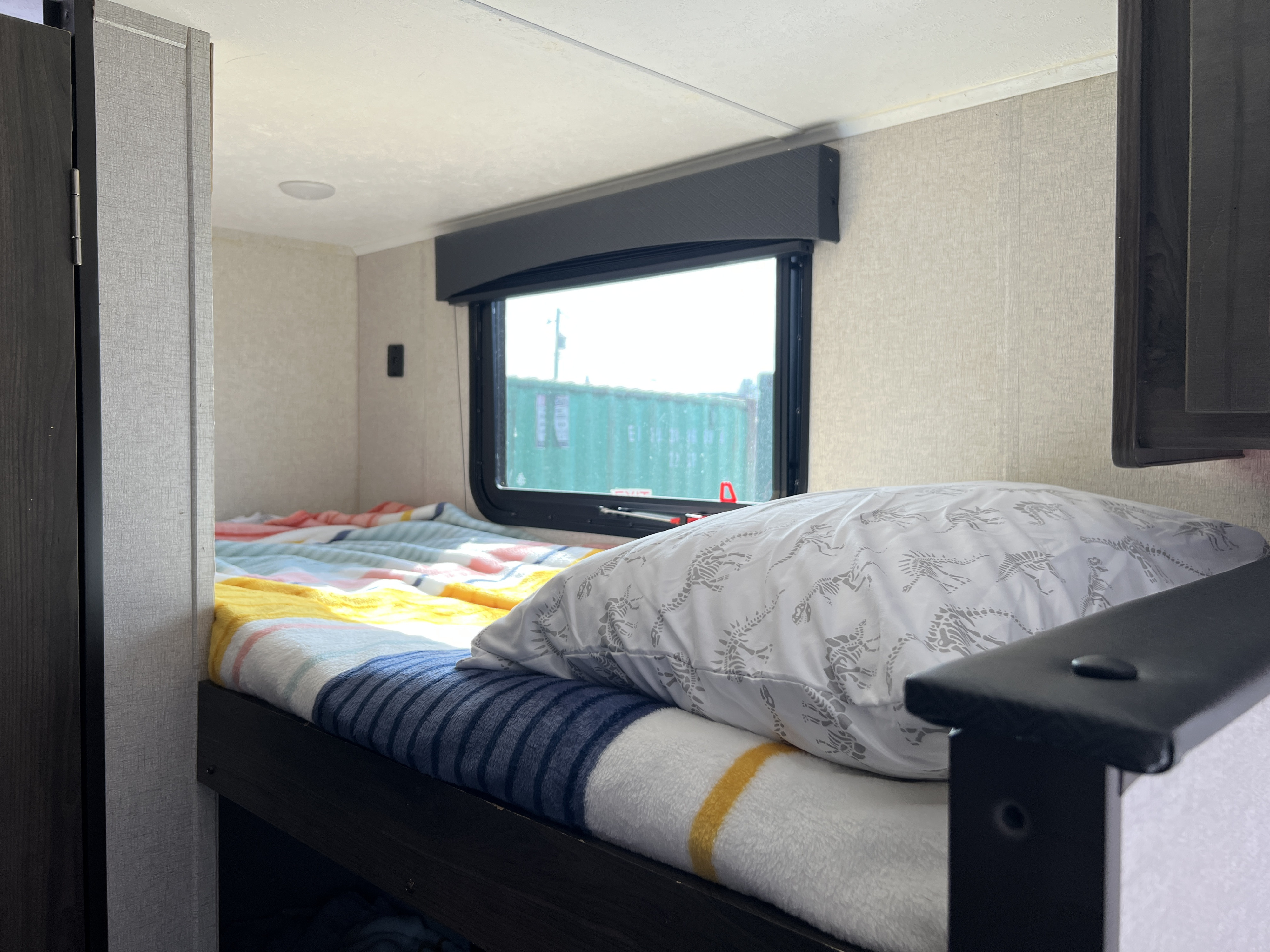 Cozy RV rental bunk bed with striped blankets and a dinosaur pillow. A window reveals a green shipping container outside.. Clipper Clipper Trailer 2021