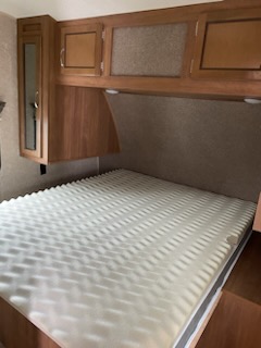 Cozy RV rental bedroom featuring a comfortable bed with a white foam topper, overhead wooden cabinets, and built-in lighting.. Coachmen Catalina 2018