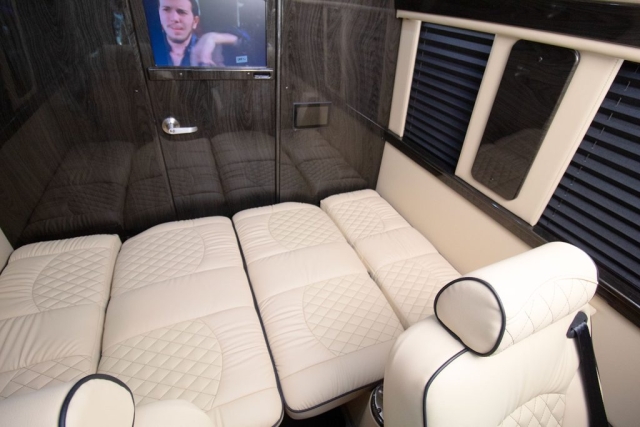 Luxury RV rental interior featuring plush cream quilted seating that converts to beds. Dark wood-paneled walls and a mounted TV offer comfort.. Ultimate Toys Ultimate Coach 2024