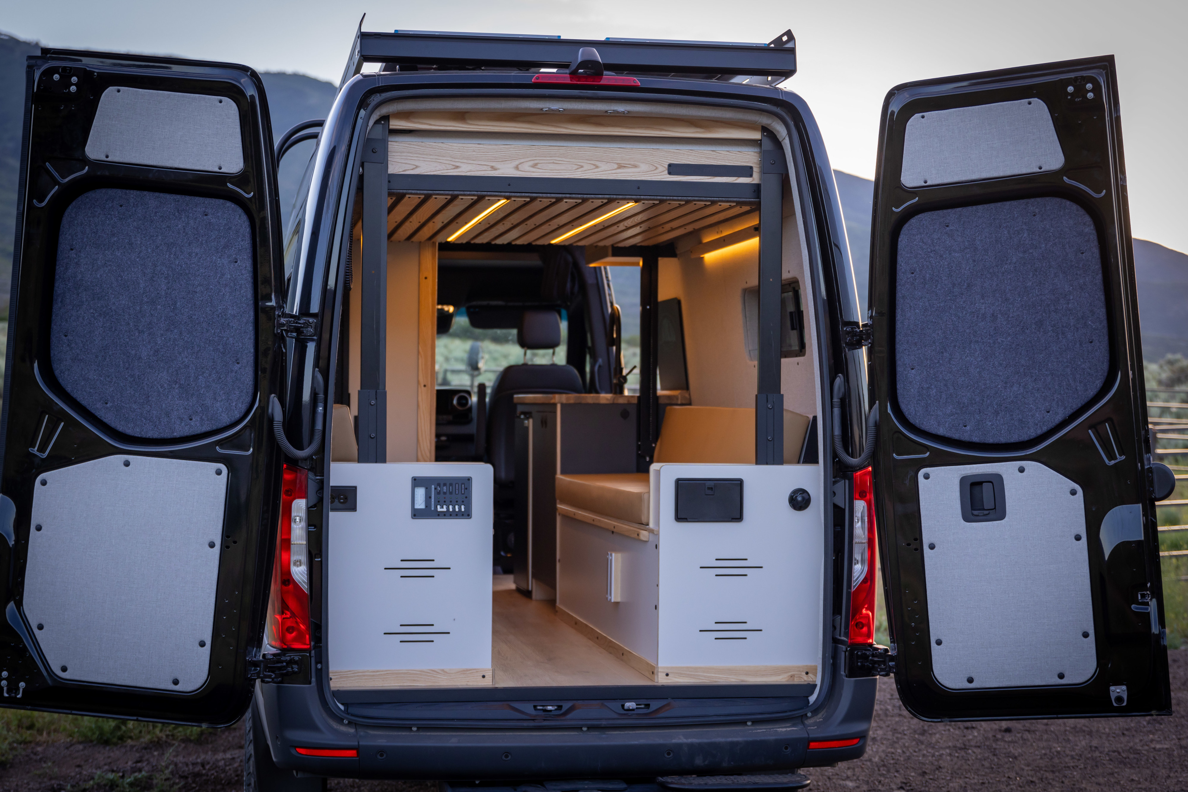 Inside an open rv rental camper van: A cozy interior with warm lighting, wood bed platform, and storage units for travel.. Mercedes Sprinter 2022