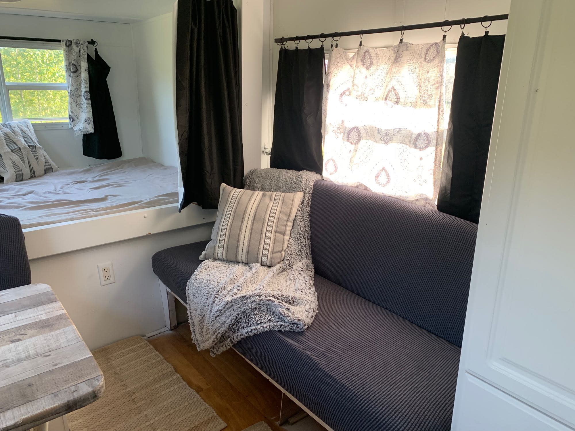 Bright RV rental interior: a comfy bed by the window, a sofa with a blanket, and wooden table.. Lite Way Outback 2003