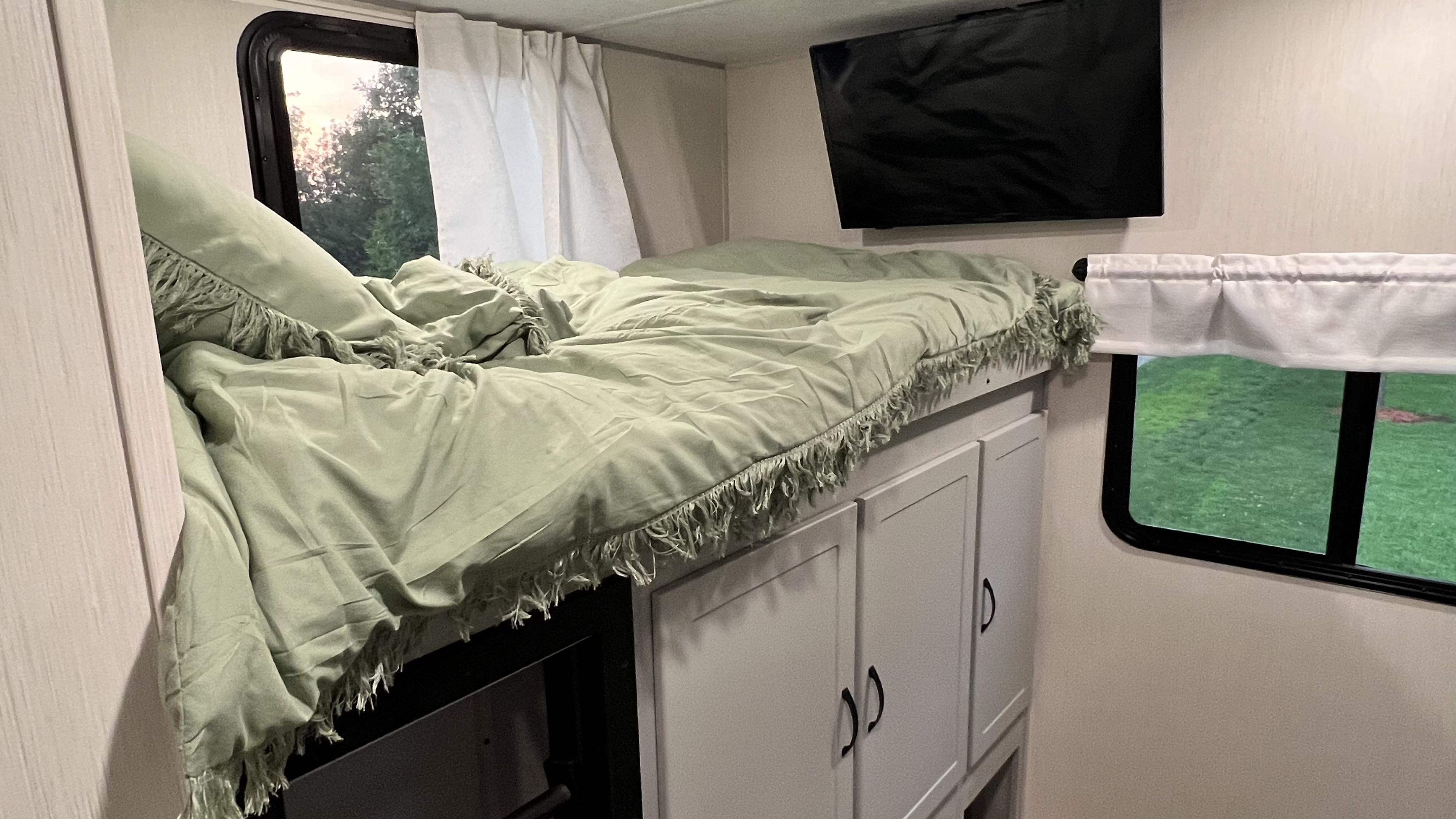 Inside a modern RV rental bedroom: a cozy bunk bed with sage green bedding, mounted TV, and built-in storage.. Forest River Vibe 2023