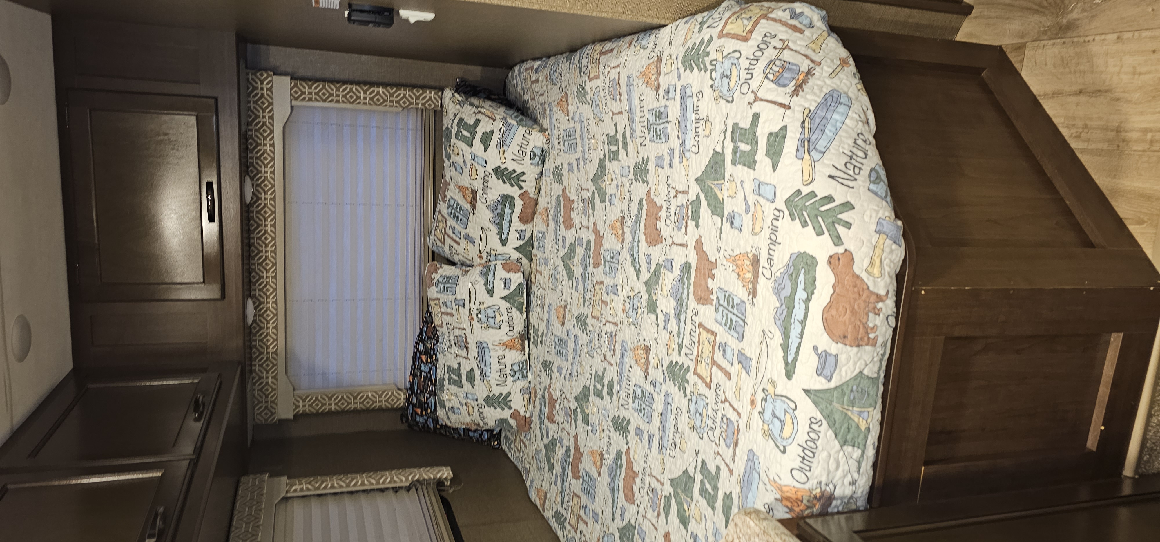 Comfortable RV rental bedroom with a fun camping-themed bedspread, dark cabinetry, and window blinds.. Thor Freedom Elite 2018