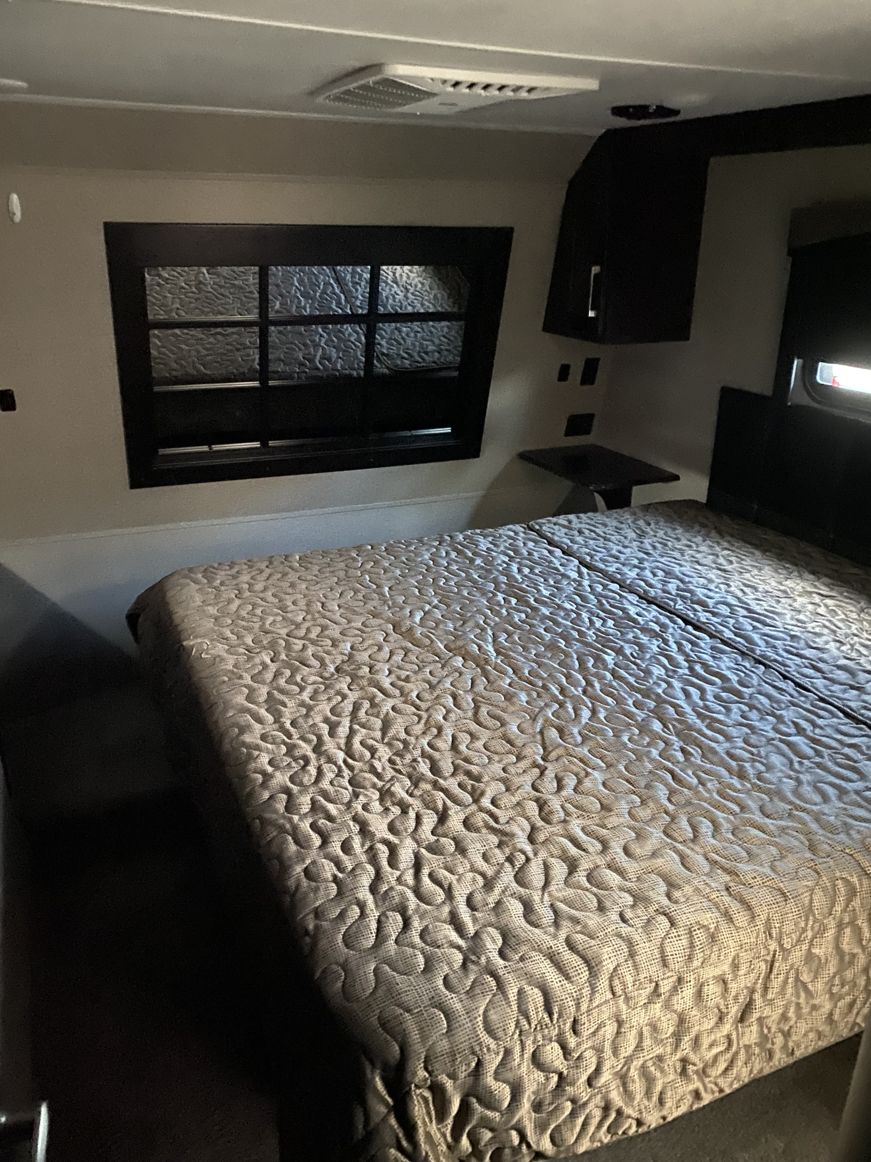 Compact RV rental bedroom featuring a textured bedspread, window, overhead cabinet, and ceiling AC unit.. Keystone Raptor 2017