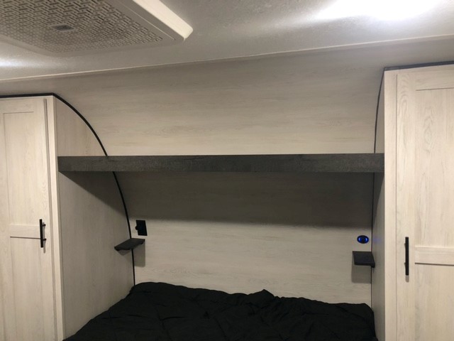 Compact RV rental bedroom showing a bed below a dark shelf, flanked by light wood cabinets. Small bedside shelves offer power and light.. KZ 301 2023