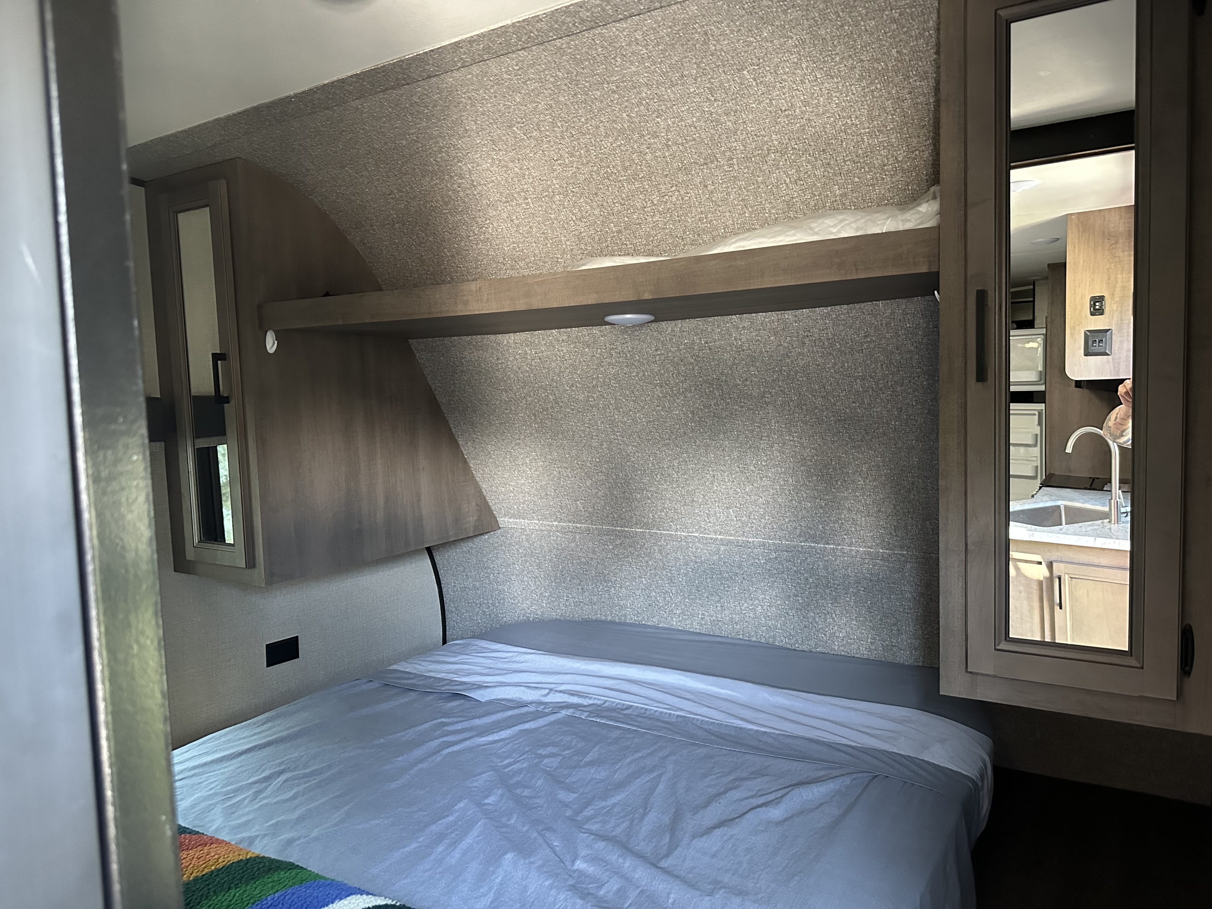 Inside an RV rental bedroom: a bed with light blue sheets, overhead shelf, and mirrored cabinet. Reflection shows a compact kitchen.. Starcraft Autumn Ridge 2022