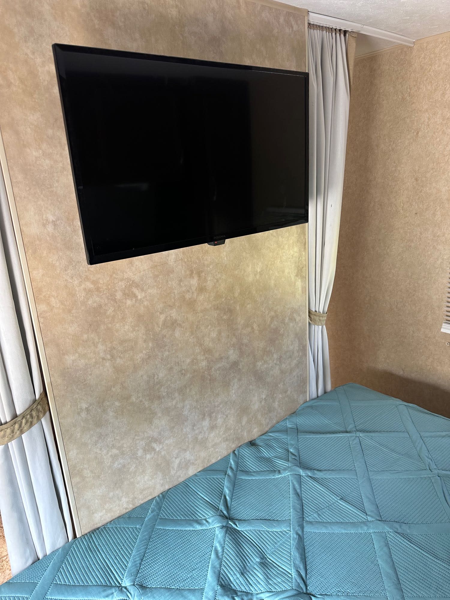 Inside an RV rental: a compact bedroom features a wall-mounted TV, quilted teal bed, and light curtains for privacy.. Jayco Jay Flight 2010