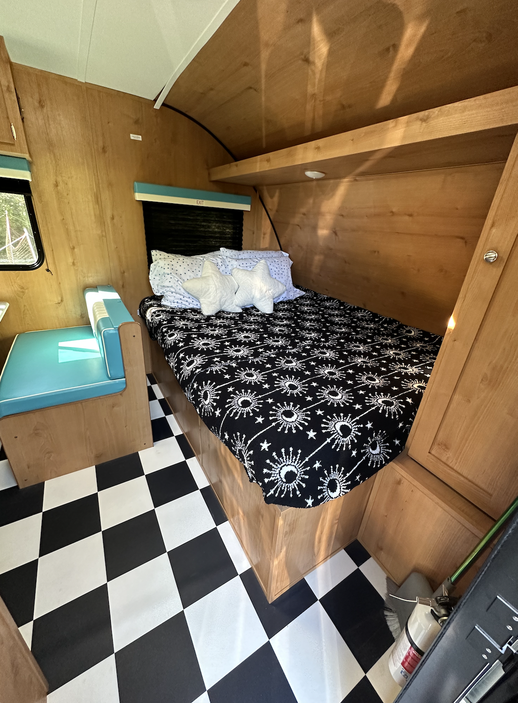 Inside a retro RV rental: bed with star pillows, teal dinette, wood paneling, and checkerboard floor. Ready for travel!. Riverside RV Retro 190BH 2023