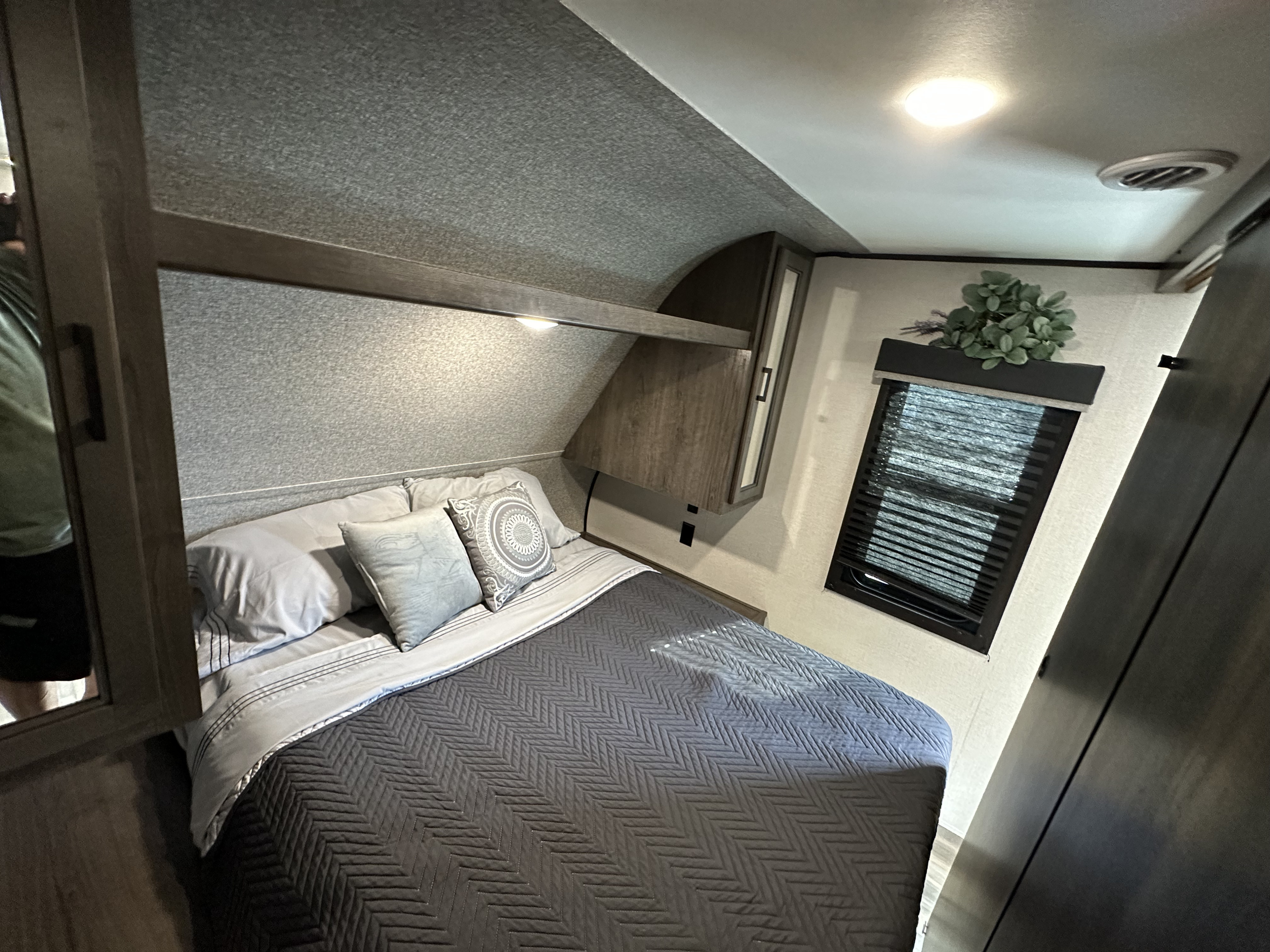 Inside an RV rental, a compact bedroom with a neatly made bed, gray linens, overhead storage, and a window with blinds.. Starcraft Autumn Ridge 2022
