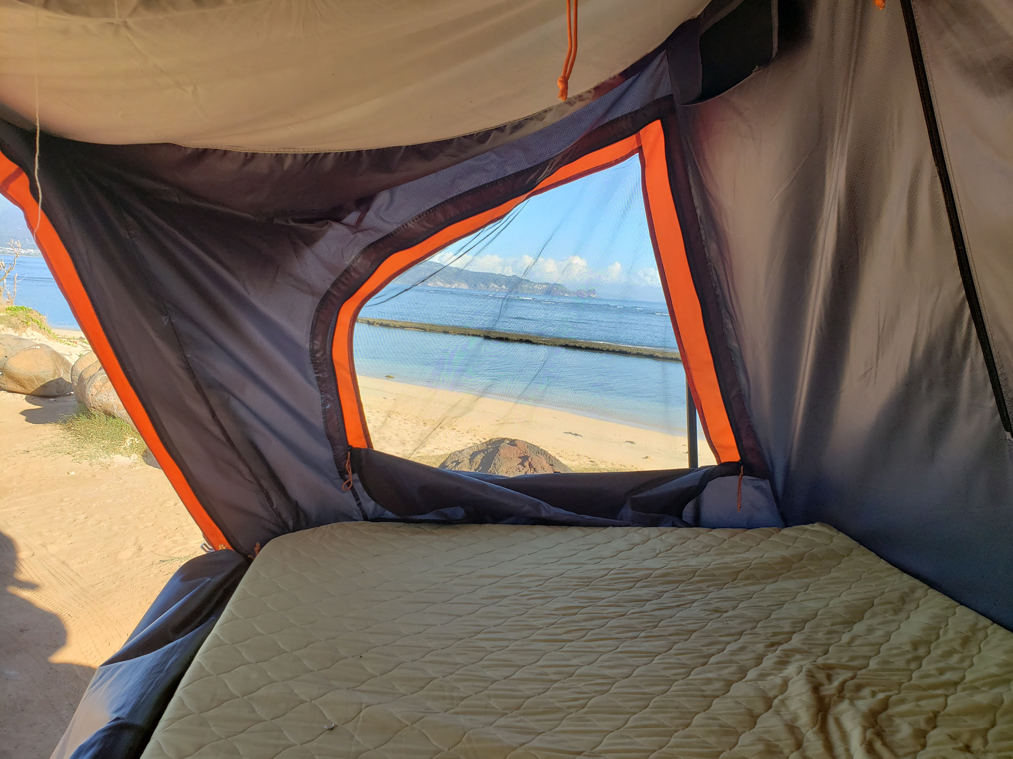 RV rental view: A quilted mattress inside a rooftop tent, looking out at a serene beach, blue ocean, and distant land.. Nissan Xterra 2012
