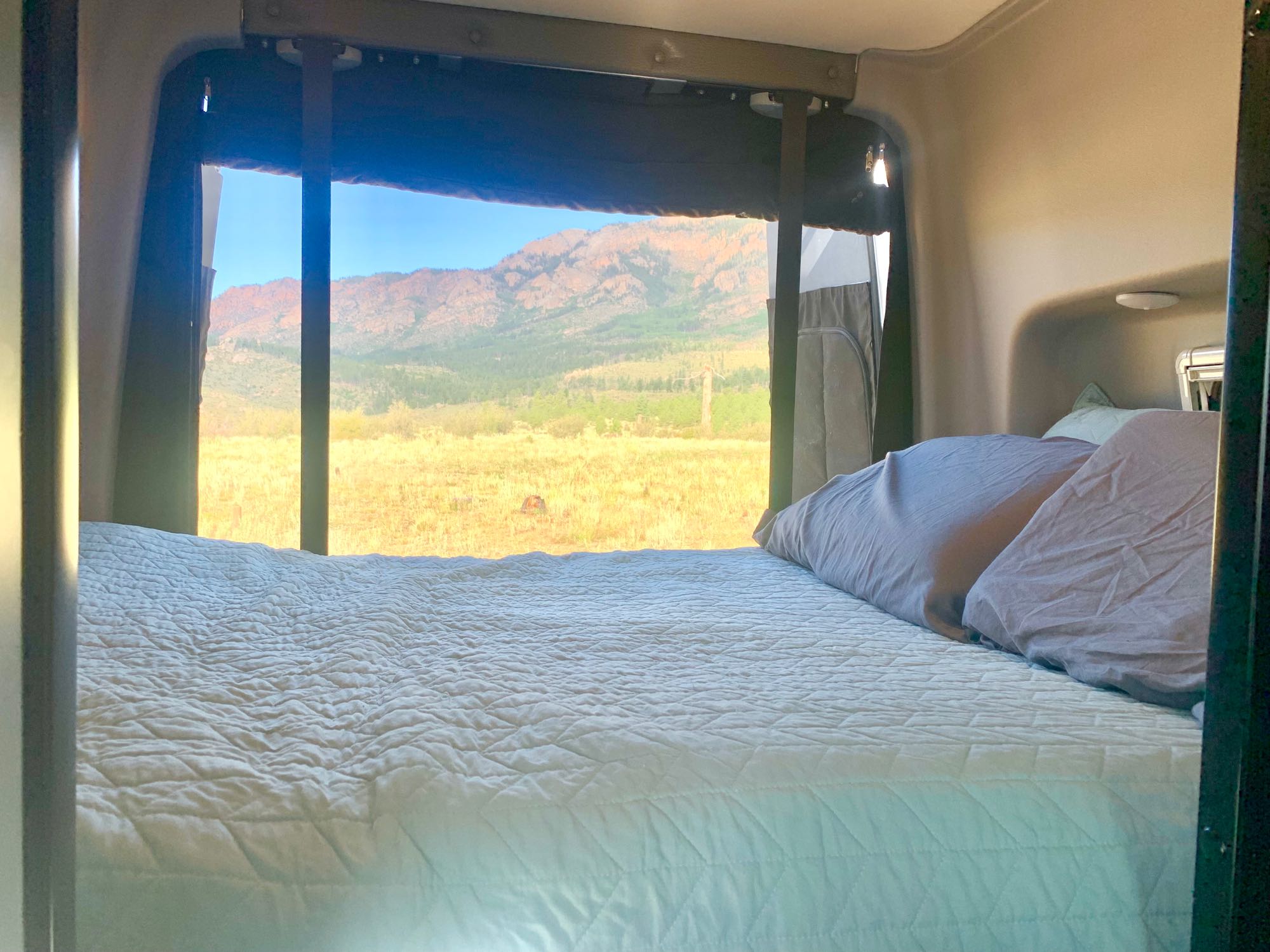 RV rental bedroom with a comfy bed and a stunning view of golden fields and mountains from the large window.. Winnebago Revel 2021
