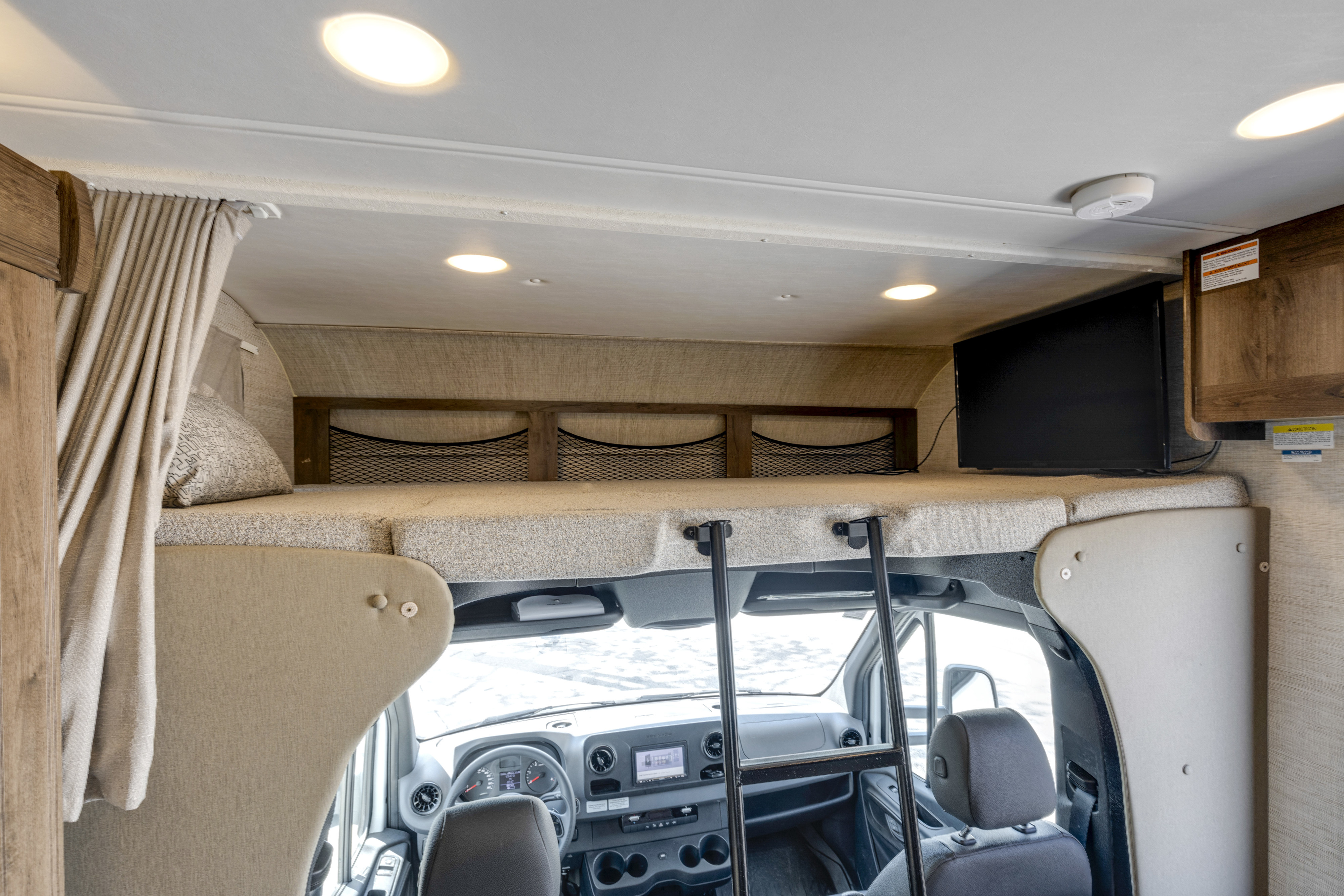 Interior of an RV rental showing the cab-over bunk with bed, safety netting, and TV, overlooking the front cabin seats.. Jayco Melbourne 2022