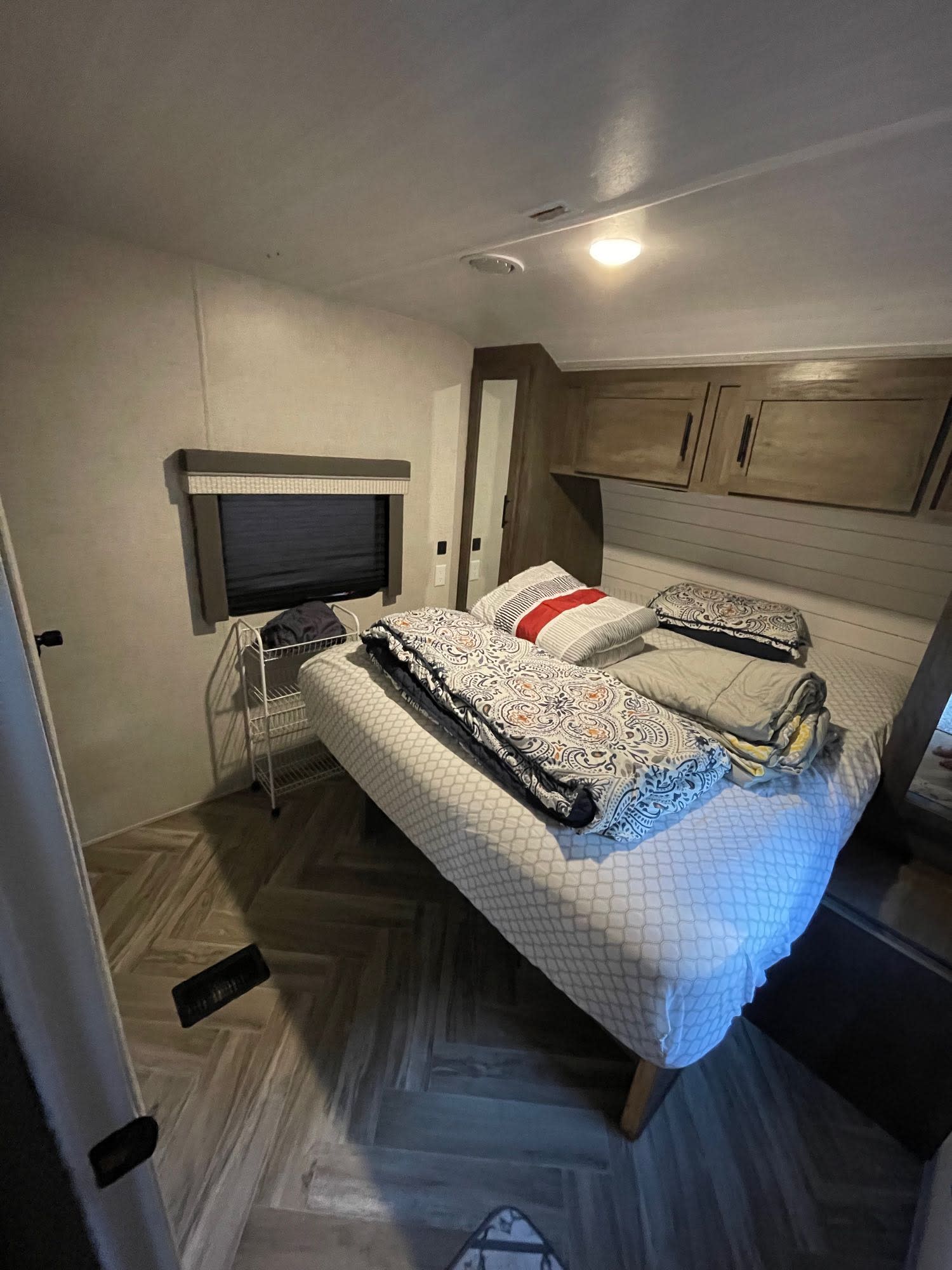 Cozy RV rental bedroom featuring a compact bed, overhead storage cabinets, a window with blinds, and wood-look flooring.. Forest River Wildwood 2021