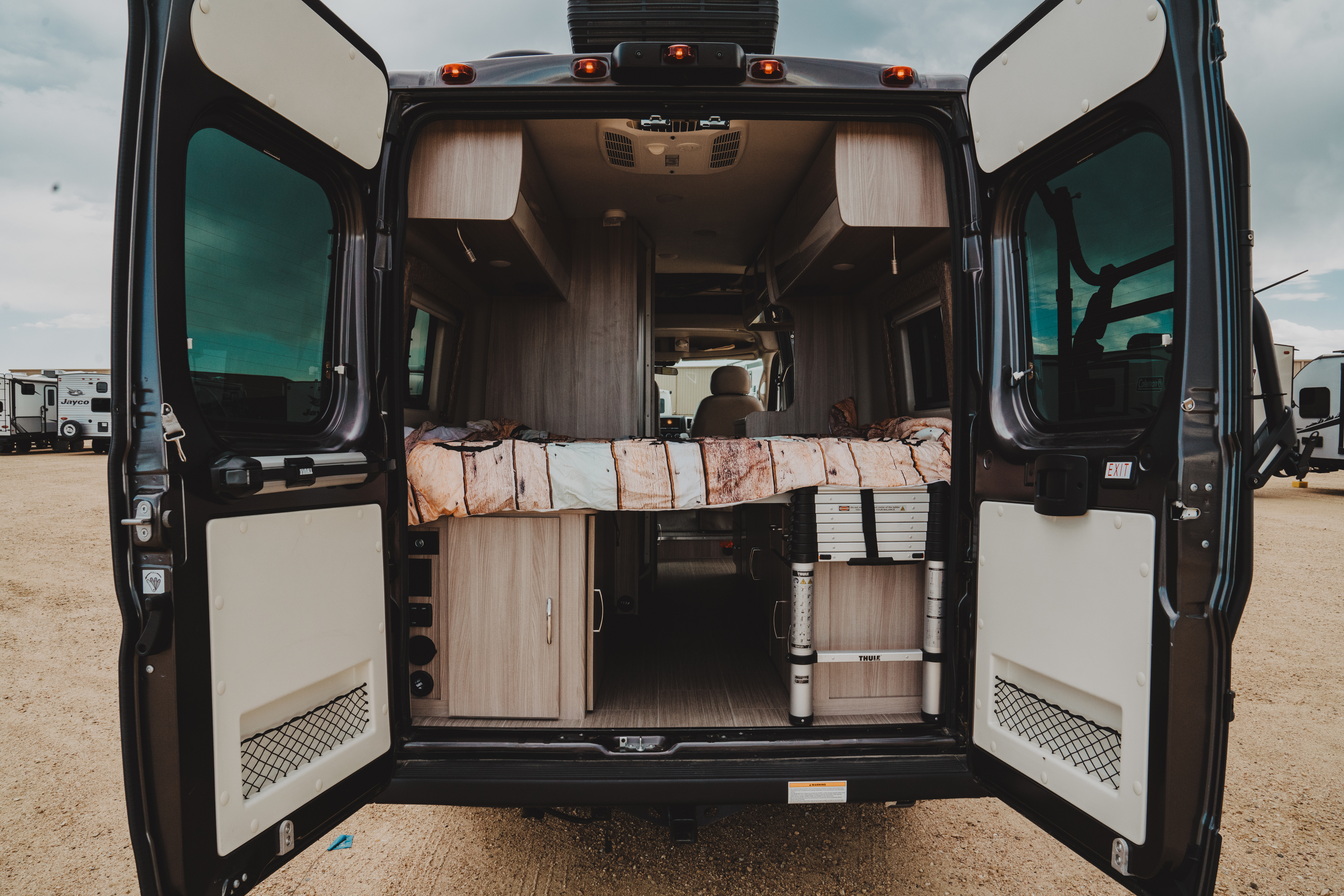 View into a modern rv rental camper van with open rear doors, showcasing its comfortable bed and organized interior.. Thor Motor Coach sequence 2021
