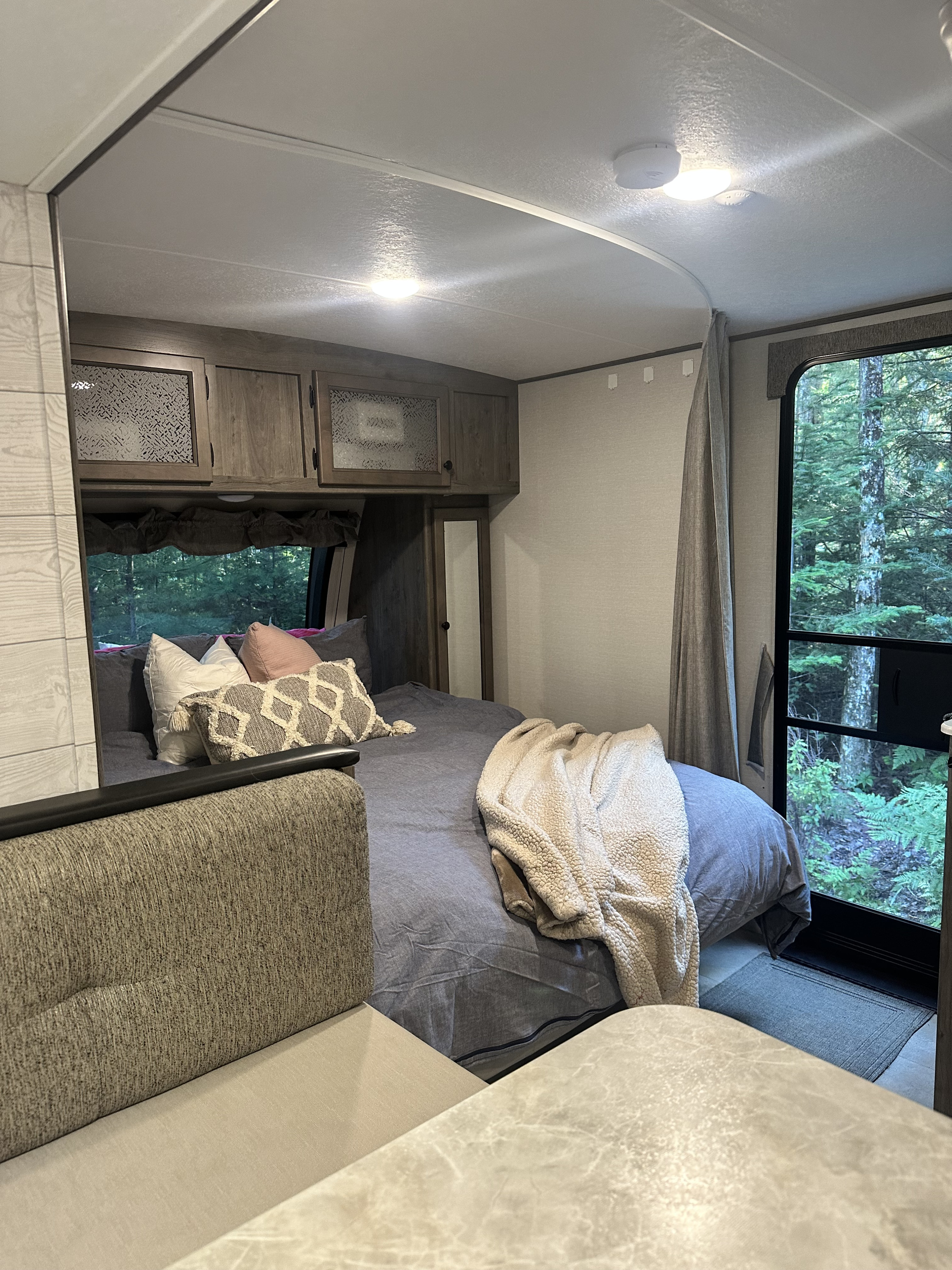 Cozy RV rental interior showcasing a bedroom with a comfy bed, overhead cabinets, and a large window with a lush forest view.. Coachmen Apex Nano 2021