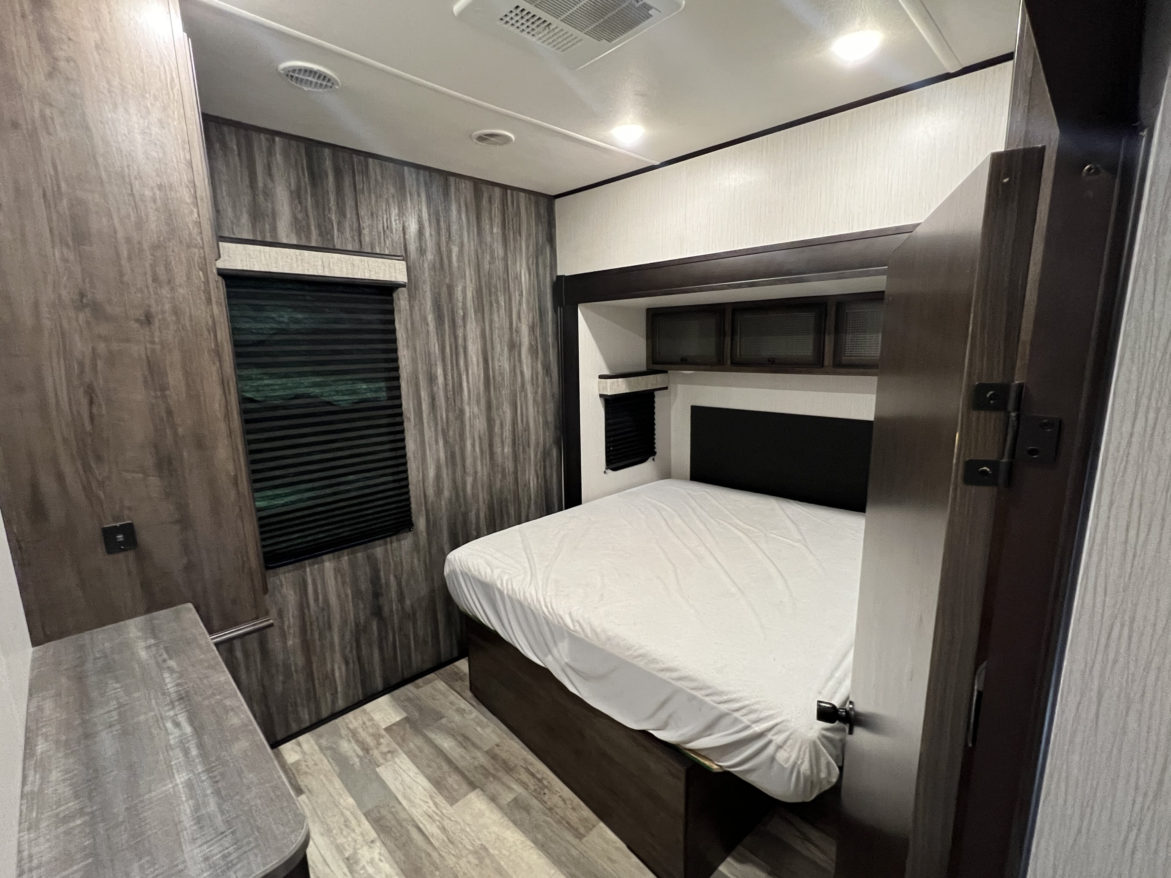 RV rental bedroom: a compact space with a bed, wood-paneled walls, window with blinds, and overhead storage cabinets.. Heartland RVs Milestone 2020