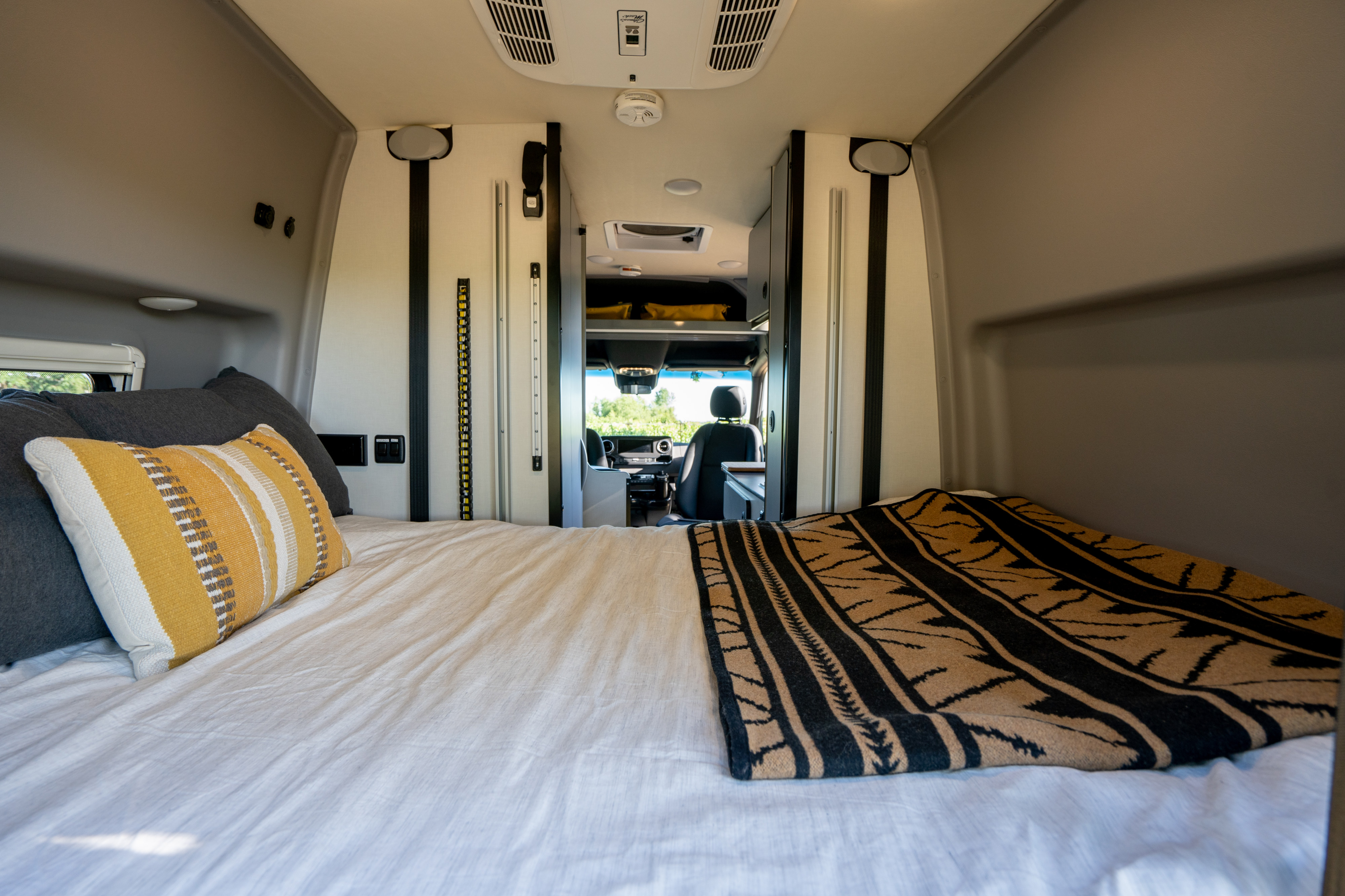 Interior of an RV rental, showing a cozy bed with pillows and a blanket, looking forward towards the driver's seat.. Winnebago Revel 2023