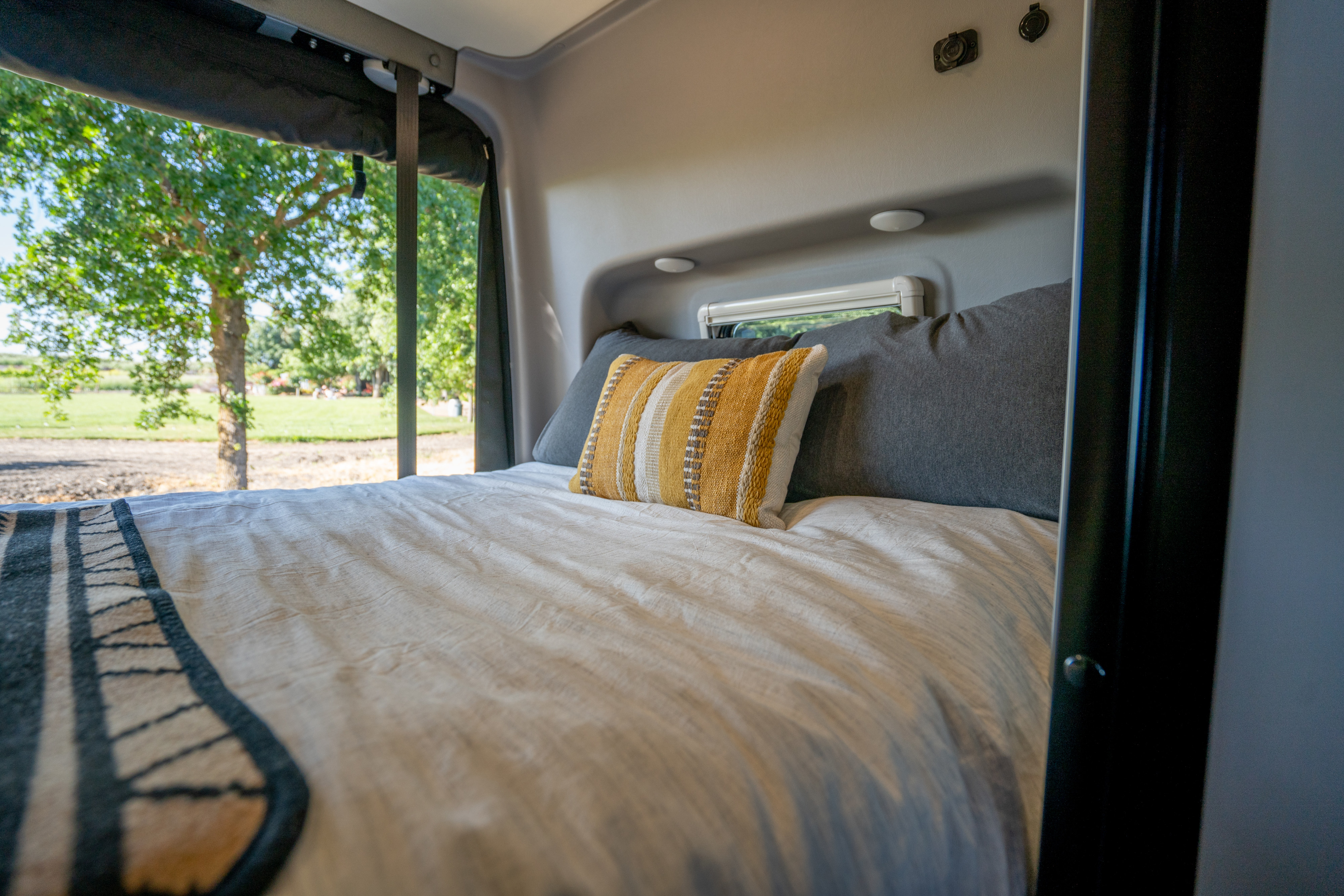 Cozy RV rental bedroom featuring a comfortable bed with a striped pillow and a serene view of nature outside the window.. Winnebago Revel 2023