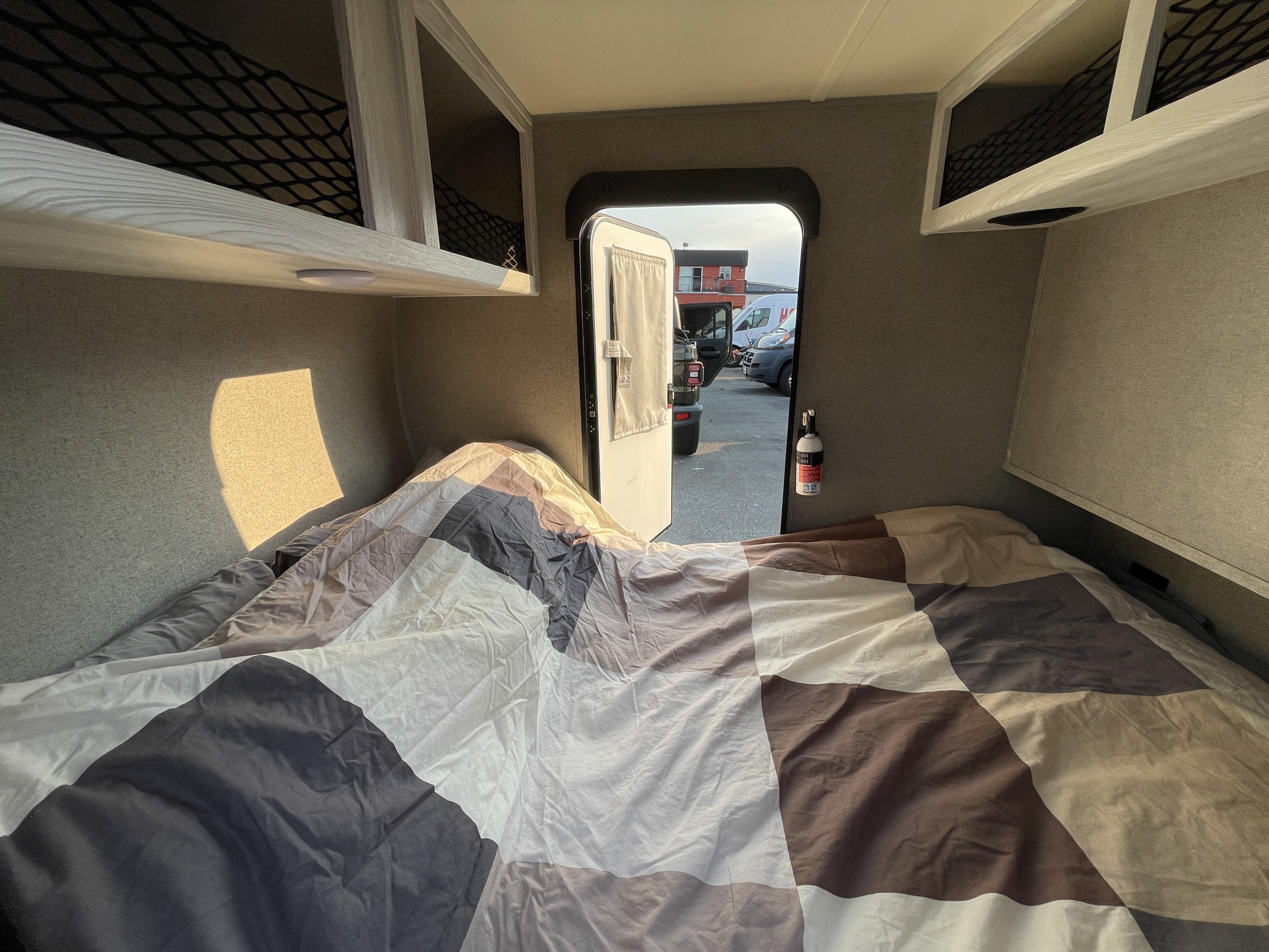 Cozy RV rental bed with checkered blanket, overhead storage, and an open door viewing a parking lot.. Forest River Rockwood Geo Pro 2018