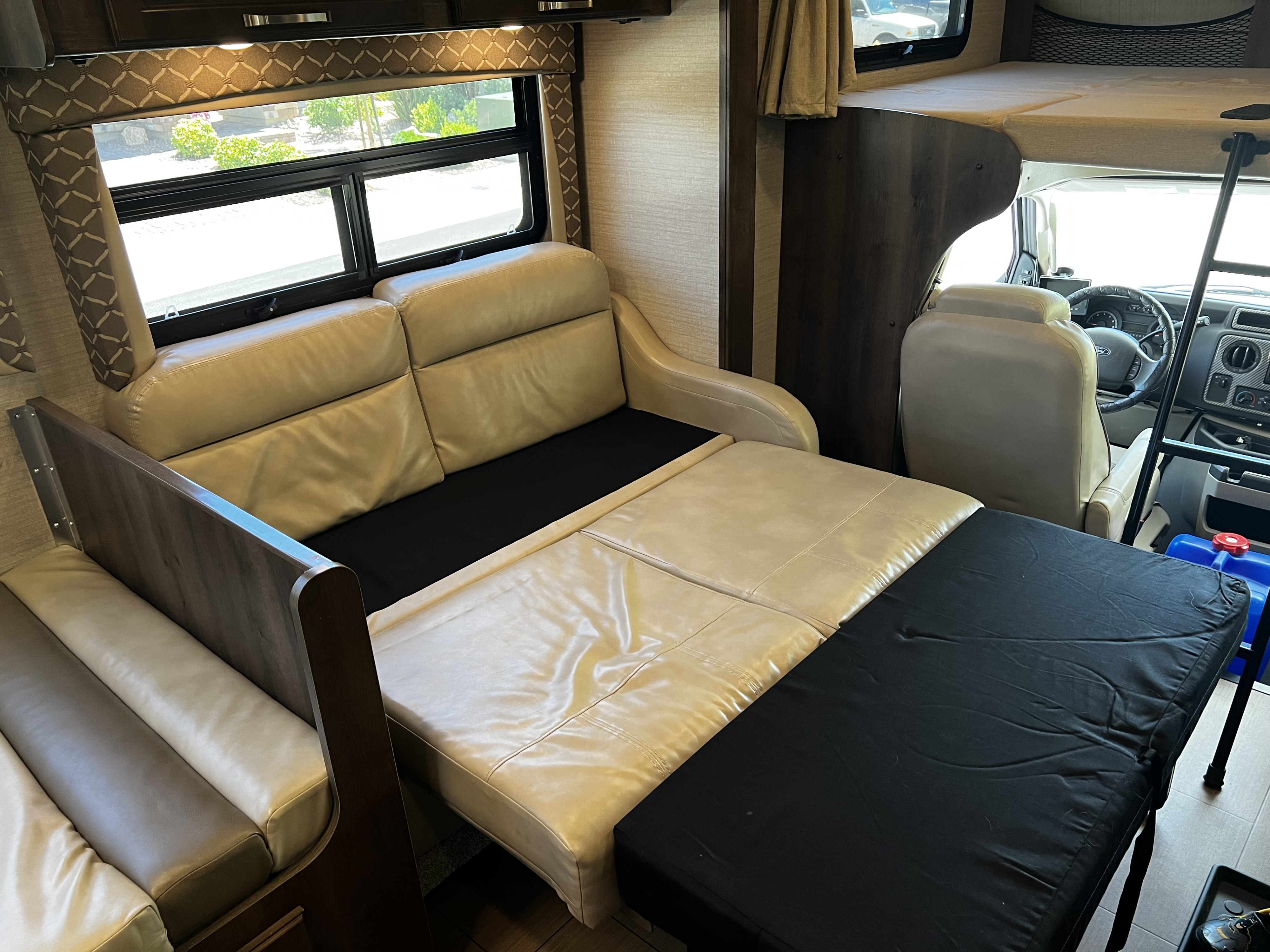 RV rental interior: A beige sofa converts to a bed, with an overhead bunk, driver's cab, and window view.. Jayco Greyhawk 2018