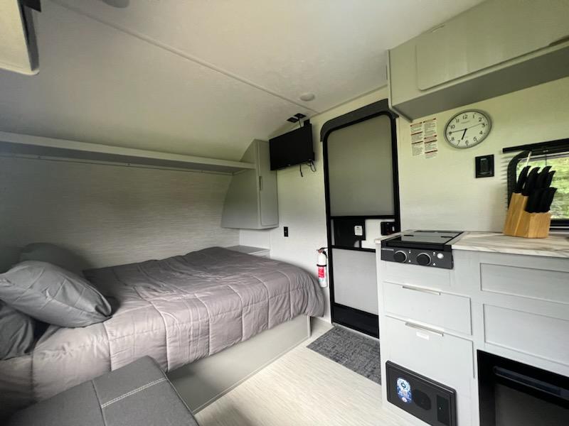 Inside this modern rv rental: a cozy grey bed, kitchen with stovetop & knife block, and wall-mounted TV. Ready for travel.. Forest River Wildwood FSX 2023