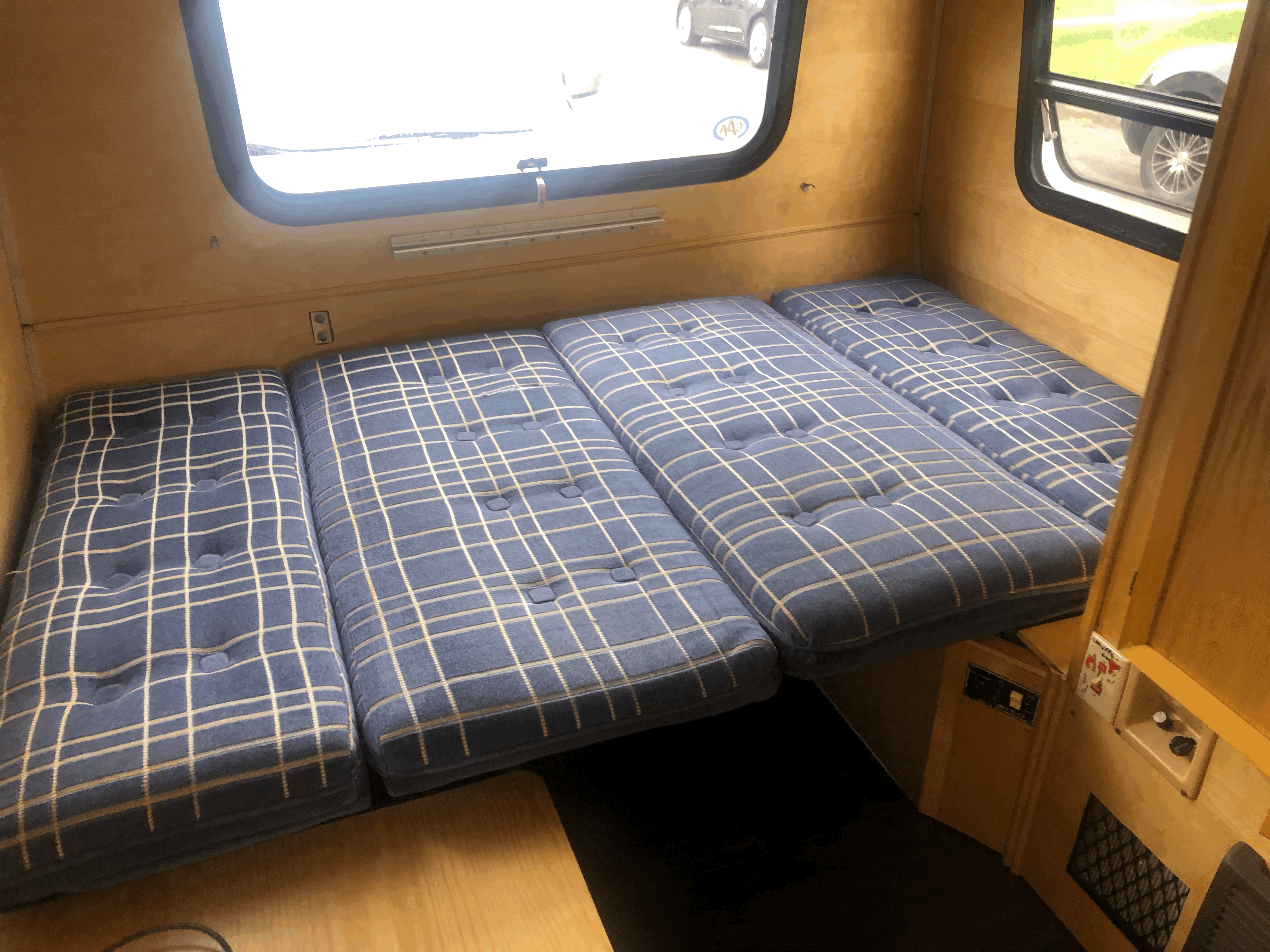 Cozy RV rental interior with blue plaid cushions arranged as a bed, light wood paneling, and windows.. Winnebago Lesharo 1985