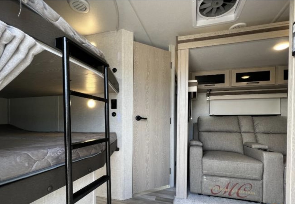RV rental interior showcasing bunk beds, a wood-grain door, and a comfortable gray seating area with overhead cabinets.. Forest River Rockwood Ultra Lite 2023