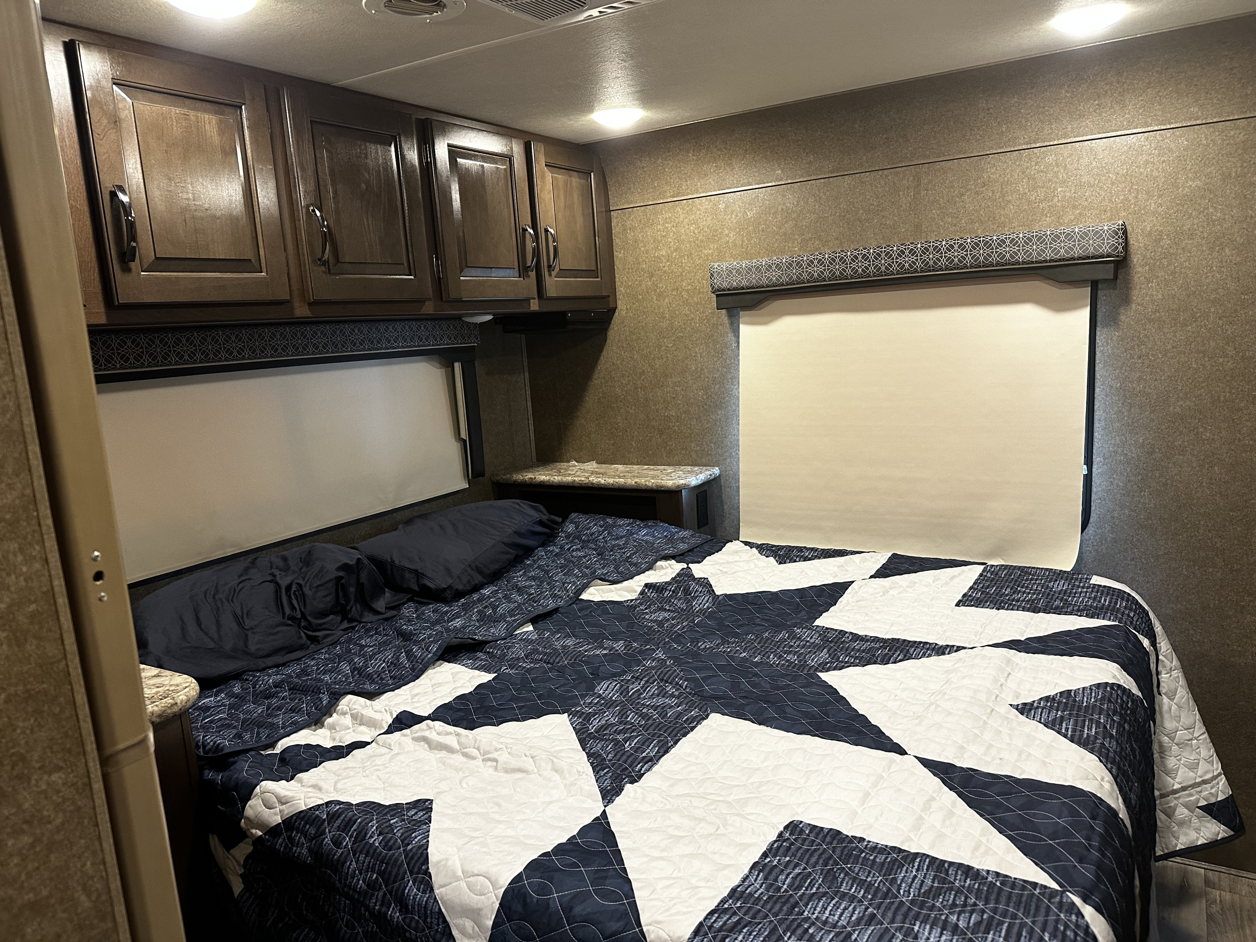 Step inside this inviting RV rental bedroom featuring a quilt-covered bed, overhead cabinets, and window privacy shades.. Thor Four Winds 2019