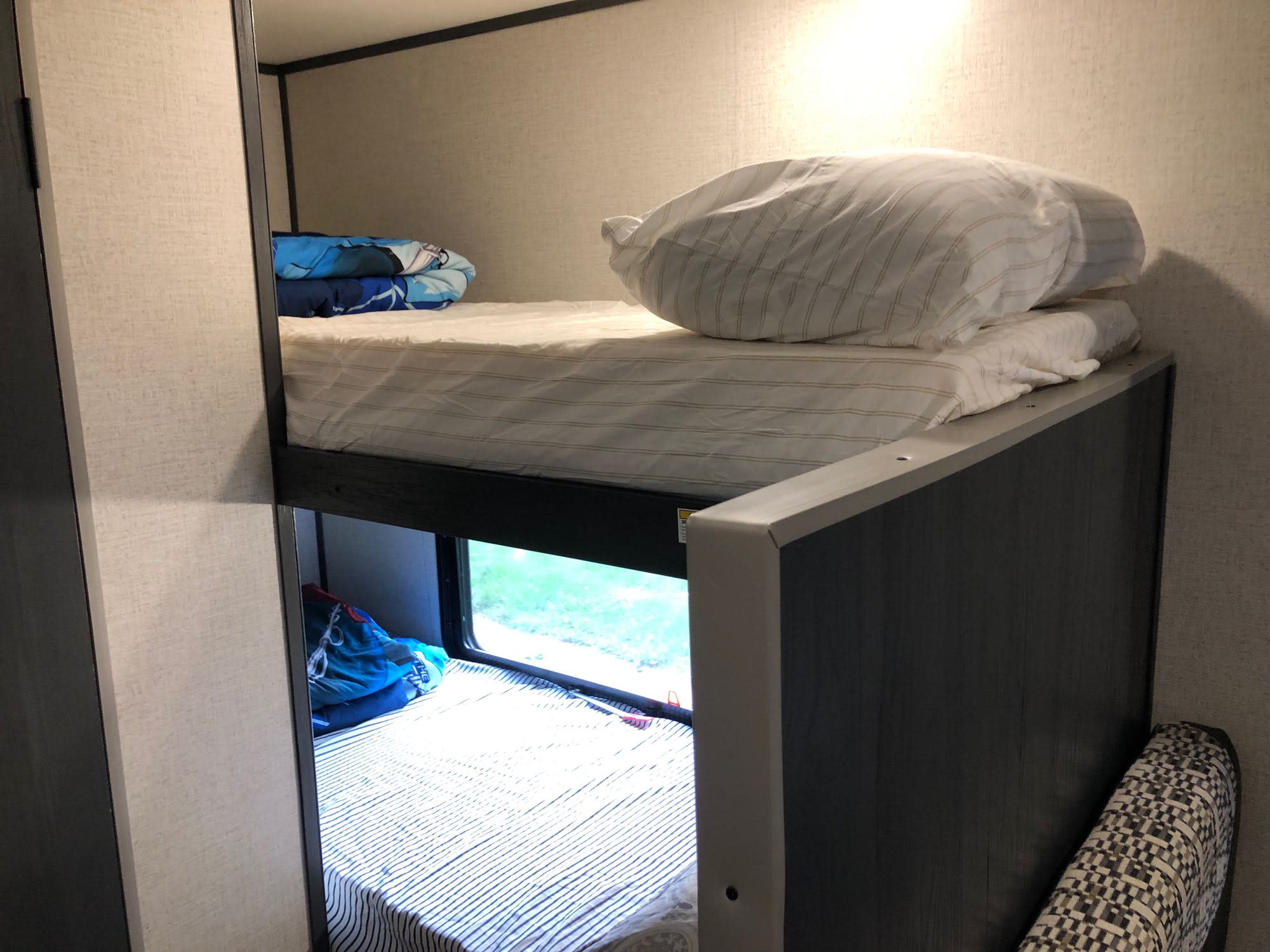 Cozy RV rental bunk beds. The top bunk features white bedding, while the bottom bunk has striped bedding and a window view.. Aspen Trail 17BH 2021
