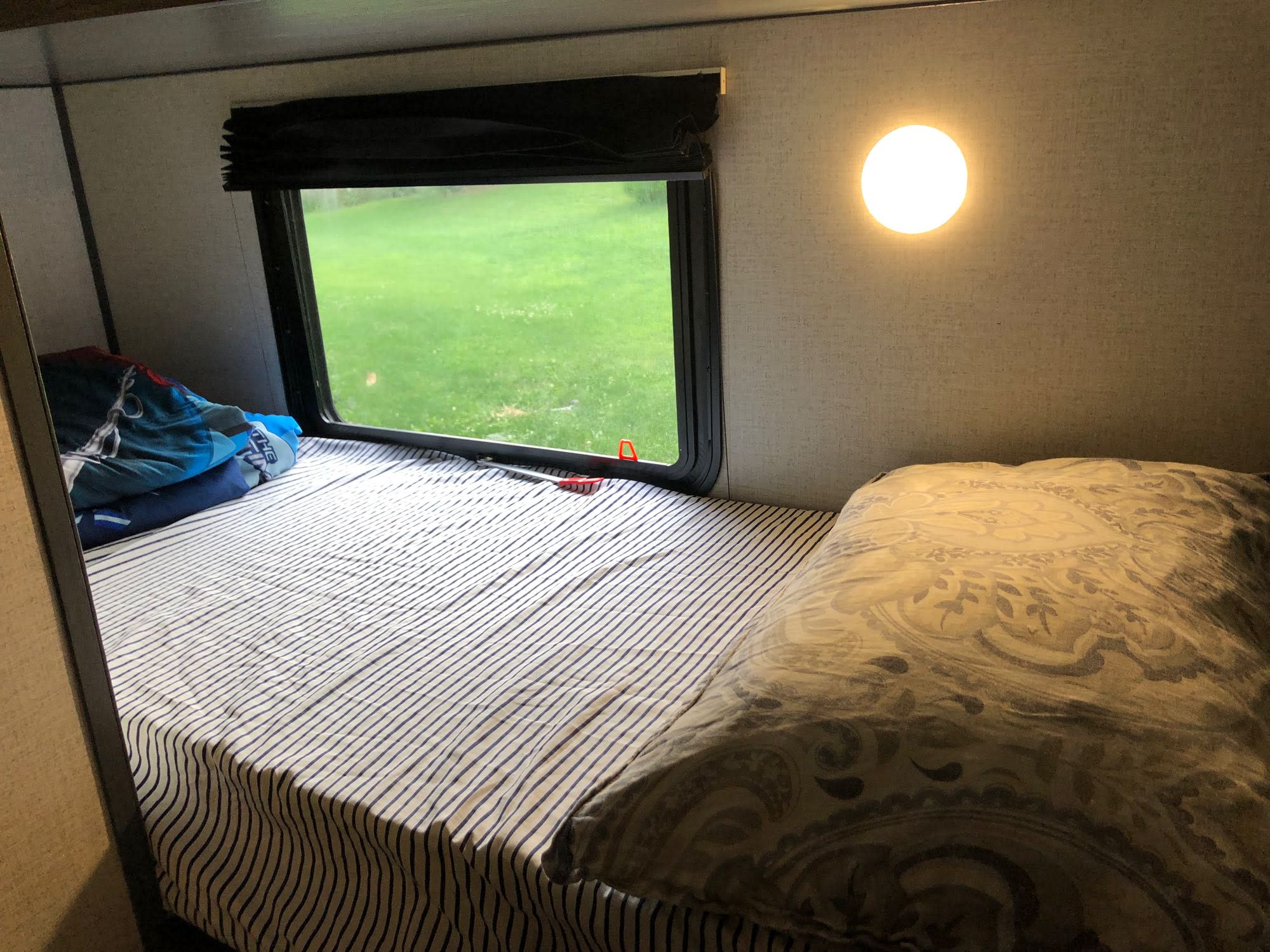 Inside a bright RV rental bedroom: a striped bed, window view of green grass, and a circular wall light. Cozy travel space.. Aspen Trail 17BH 2021