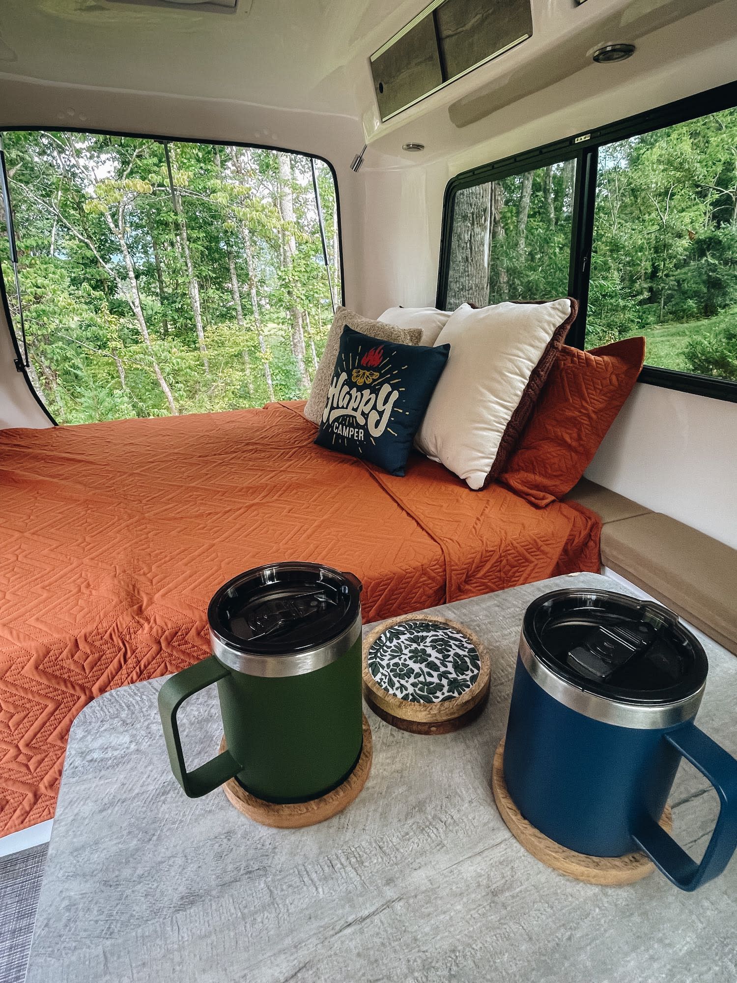 Cozy RV rental: bed with "Happy Camper" pillow, forest view, and mugs for a perfect glamping trip.. Happier Camper HC1 2022