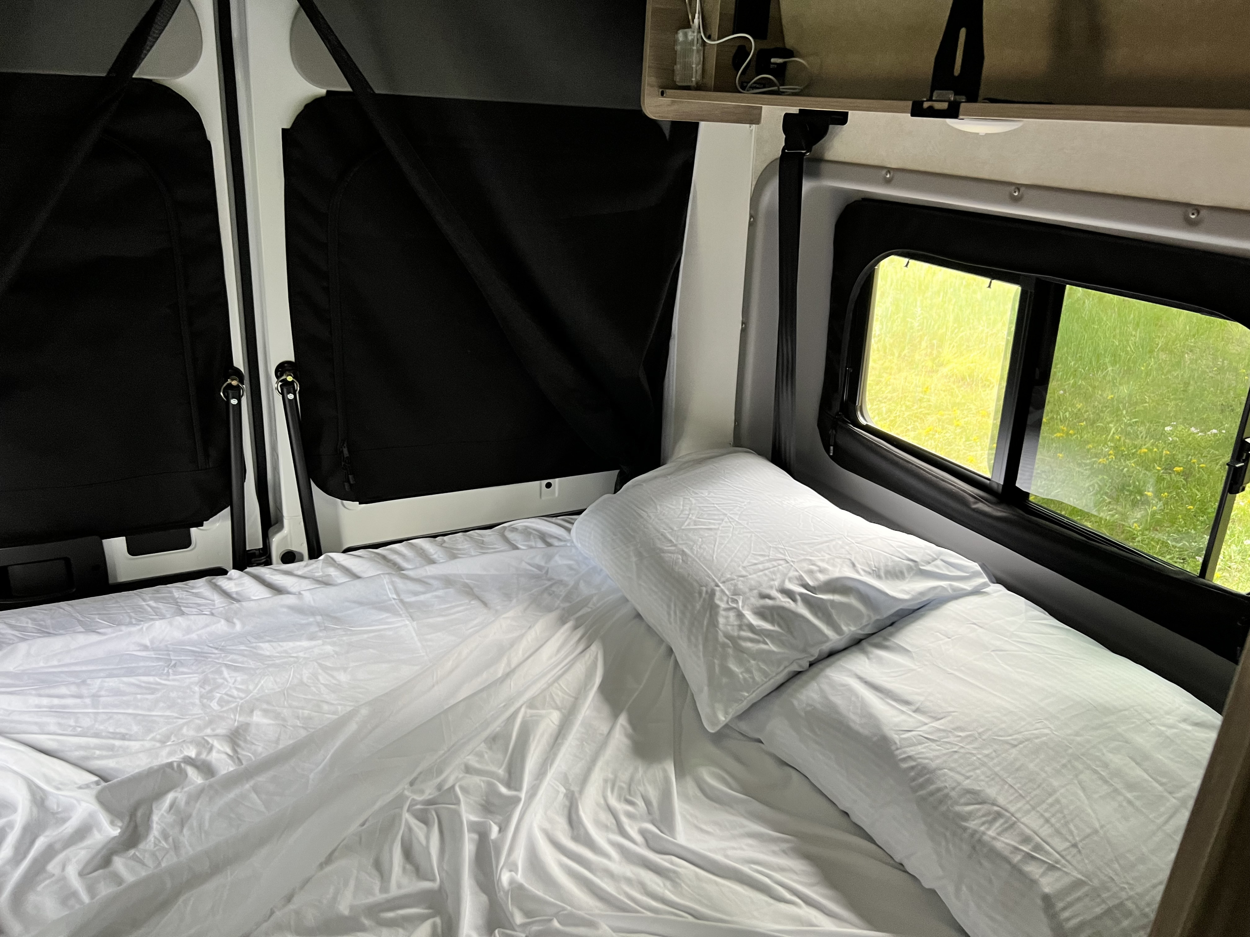 Inside a comfy RV rental: A neat bed with white sheets and pillows next to windows showing a bright green outdoor scene. Ideal for travel rest.. Winnebago Solis 2023