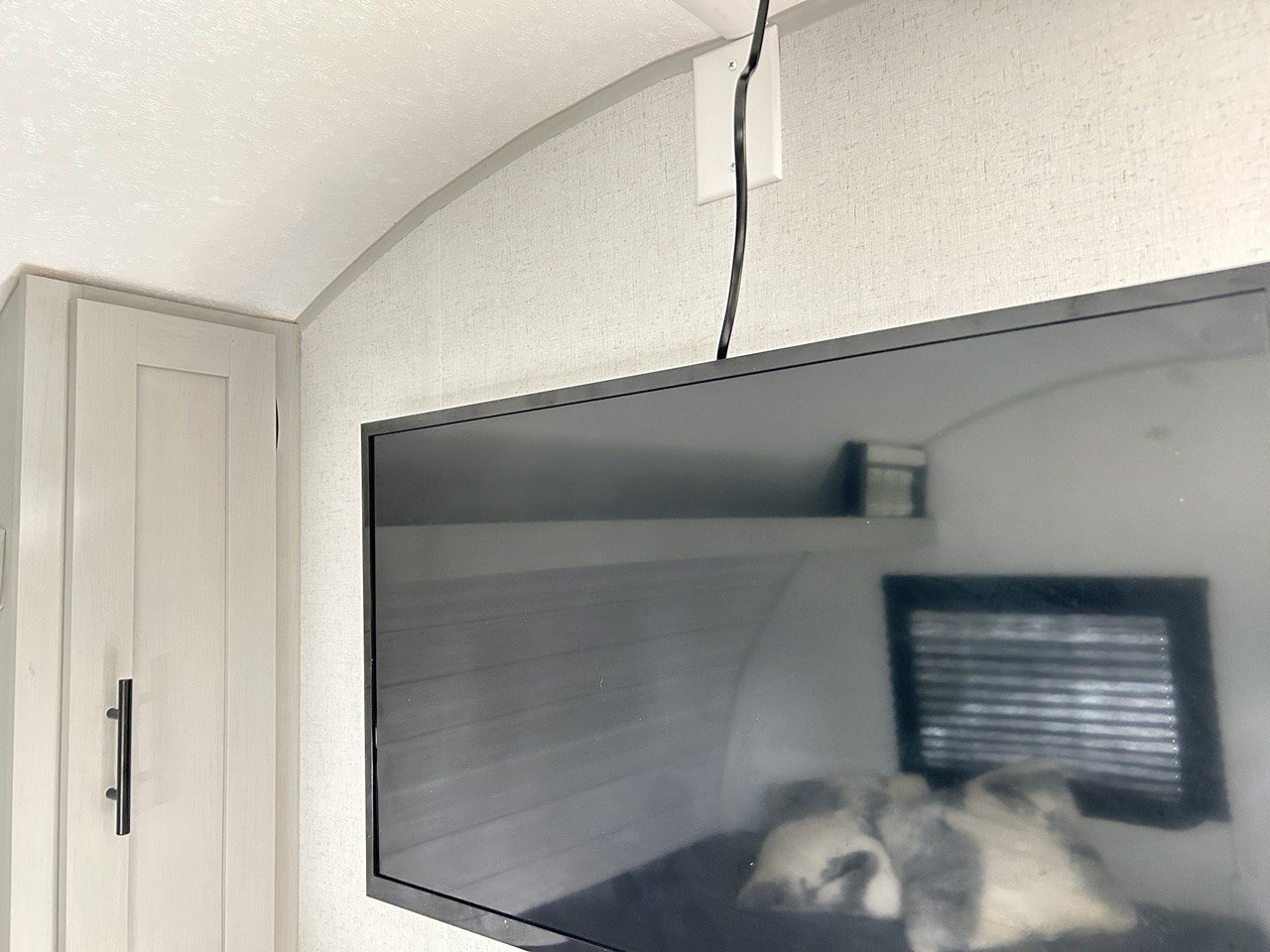 Compact RV rental interior featuring a wall-mounted TV, cable, and a light cabinet. Reflection shows a bed area.. Forest River Wildwood 2022