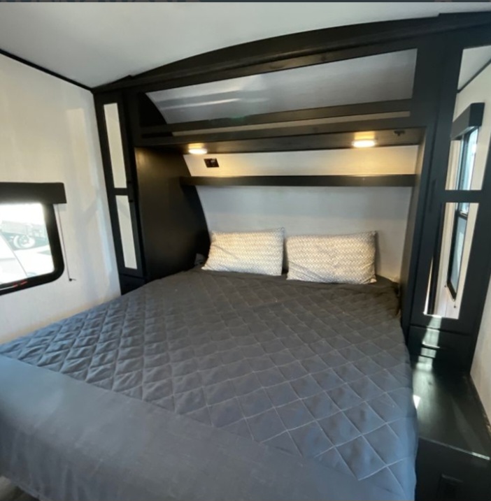 A modern RV rental bedroom featuring a comfortable bed with a dark grey comforter, overhead shelving, and convenient lighting.. Heartland RVs Fuel Toy Hauler 2022